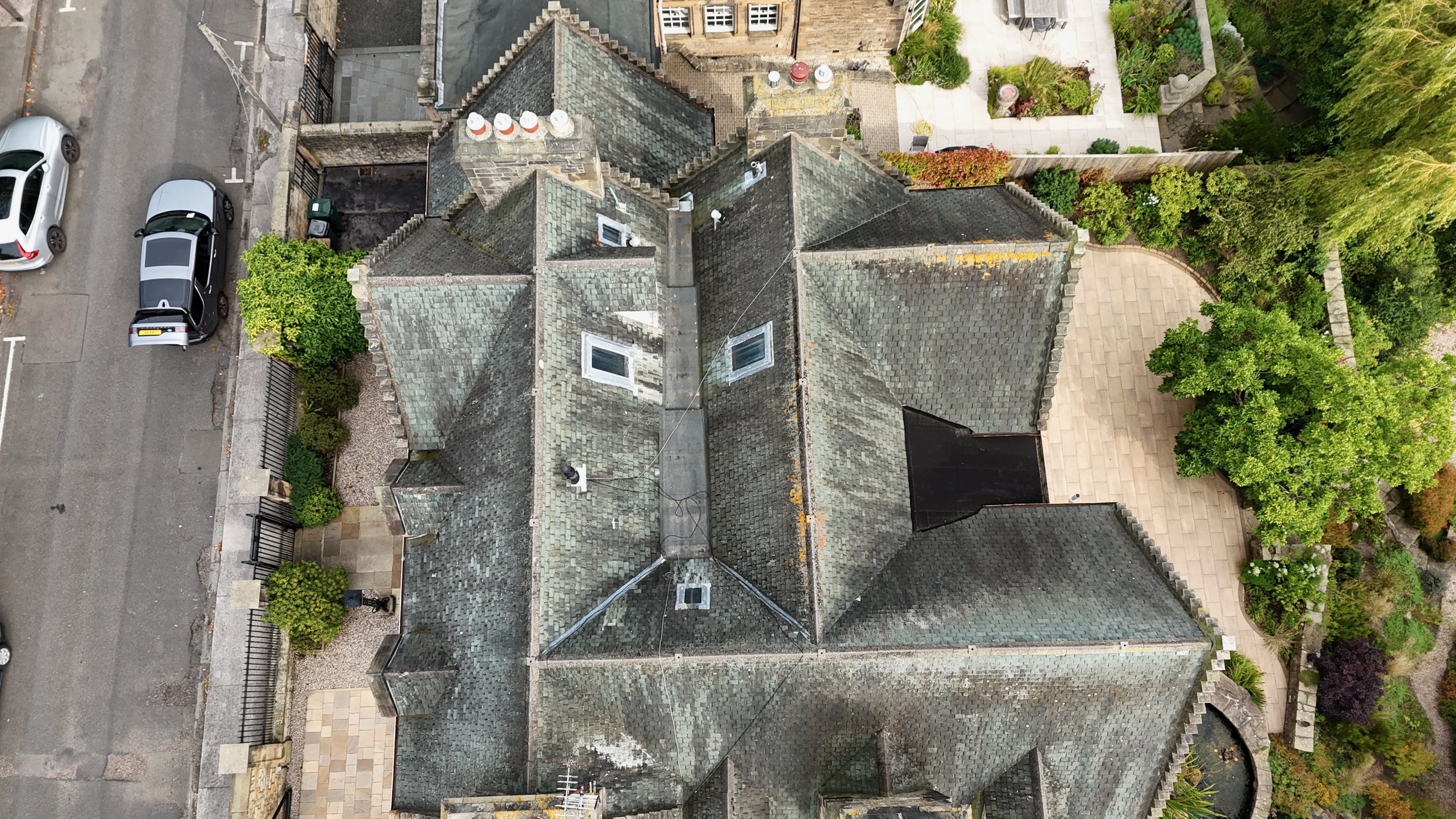 An aerial view of a gray stone building with multiple gabled roofs, surrounded by a garden, trees, and a paved walkway, with cars parked along the street.