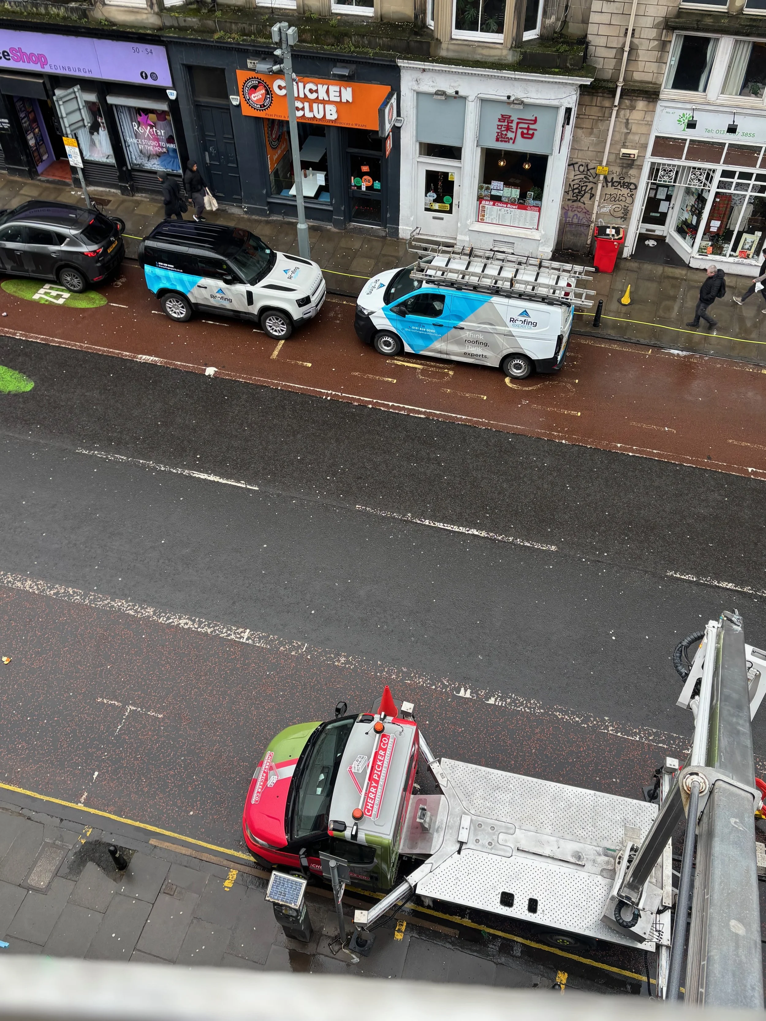 Street view showing parked cars, a construction or service vehicle with a cherry picker, storefronts including a chicken club, and pedestrians walking on the sidewalk.