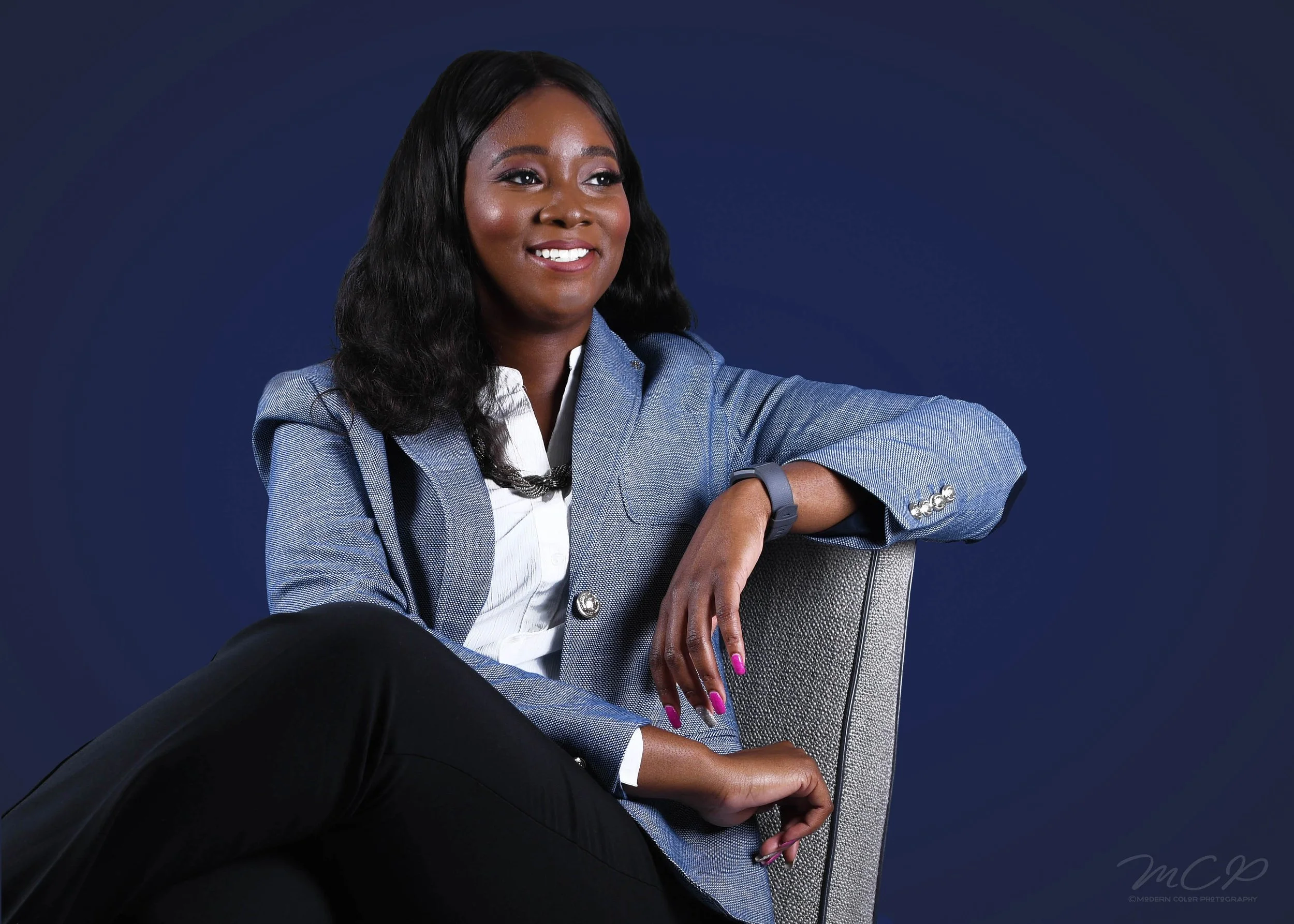 A woman with long black hair wearing a light blue blazer and black pants sitting on a gray chair against a dark blue background, smiling and looking slightly to her left.