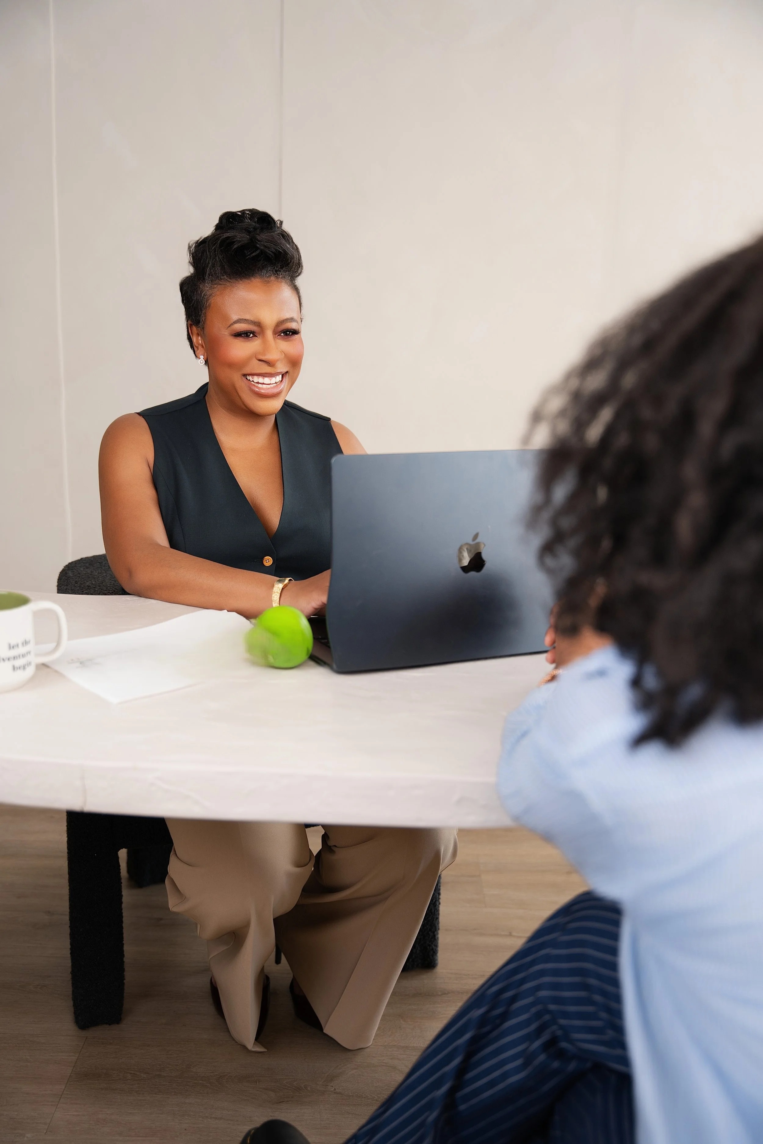 A woman with short dark hair, wearing a sleeveless black top and beige pants, sitting at a white table and smiling while looking at a person with curly hair in the foreground. On the table are a white mug, a green apple, and papers, with a laptop in front of the woman.