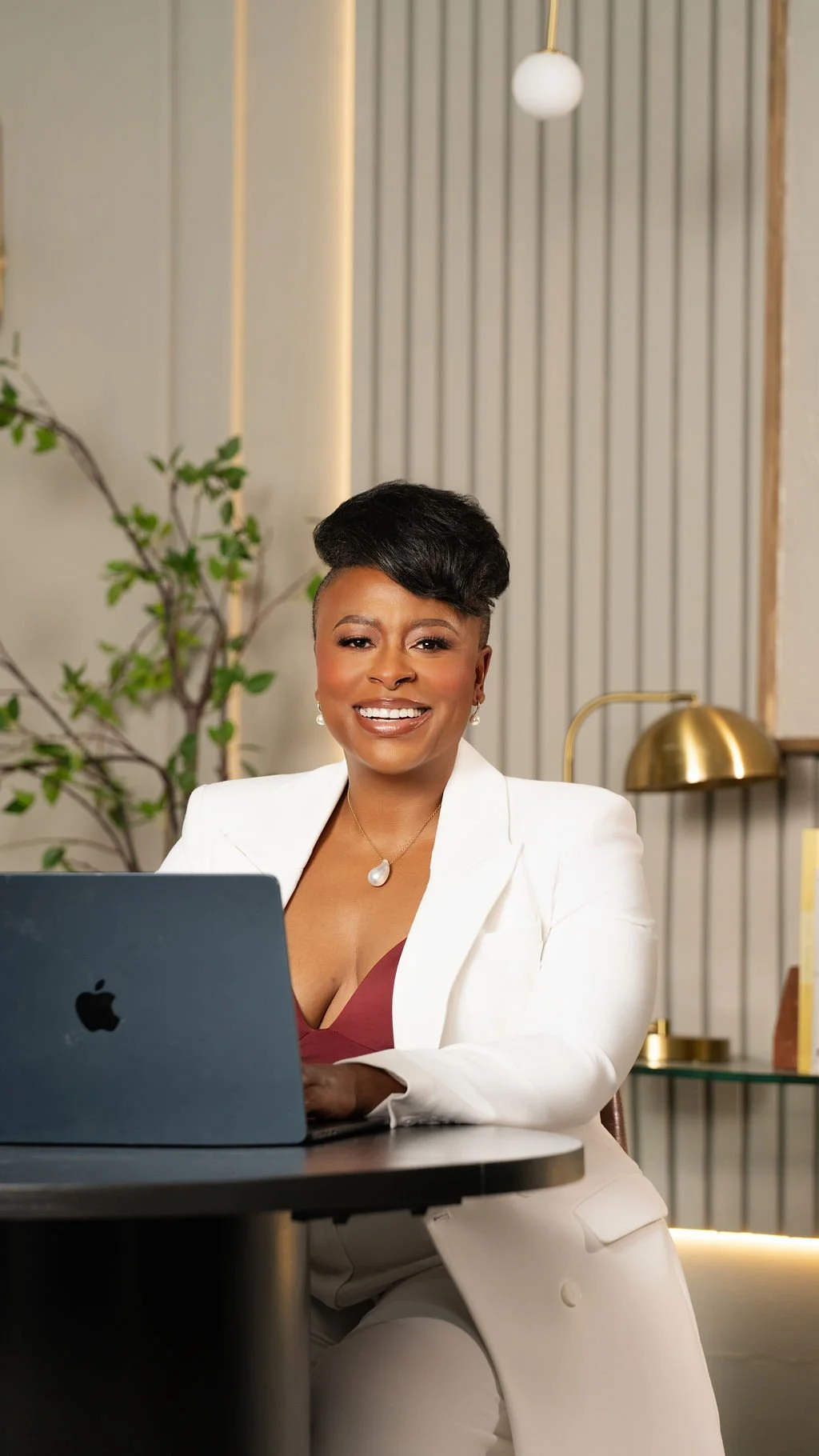 A smiling woman with short dark hair wearing a white blazer and burgundy top, sitting at a desk with an open MacBook in a modern office space.