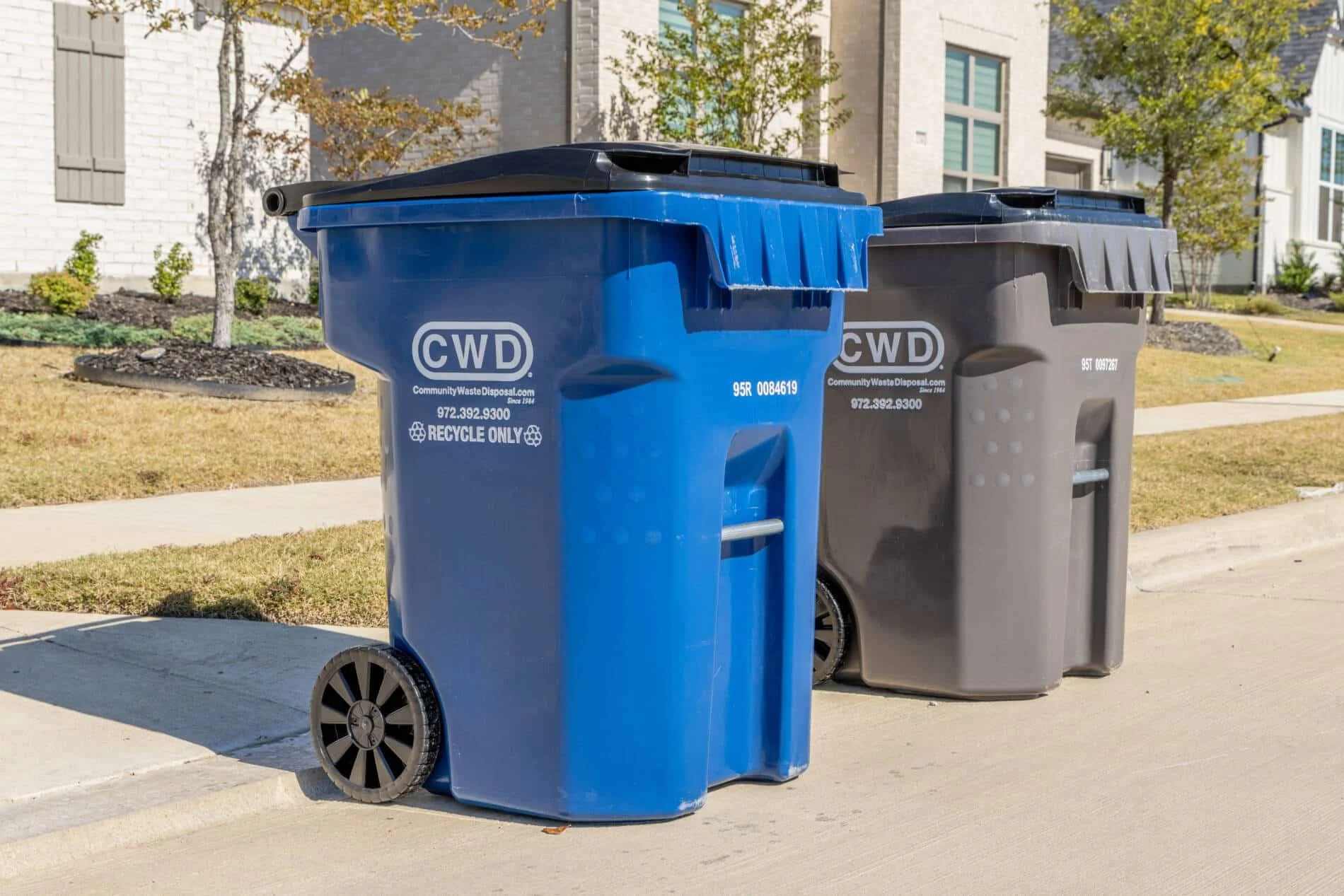 Three trash and recycling bins lined up on the sidewalk in front of houses with trees and lawns, under a clear sky.