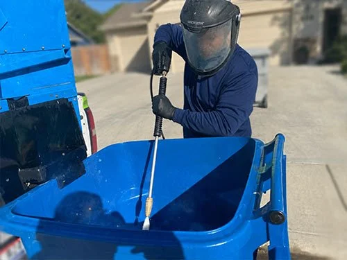 A person wearing a helmet, gloves, and a blue shirt cleaning a blue trash bin with a pressure washer in a suburban driveway.