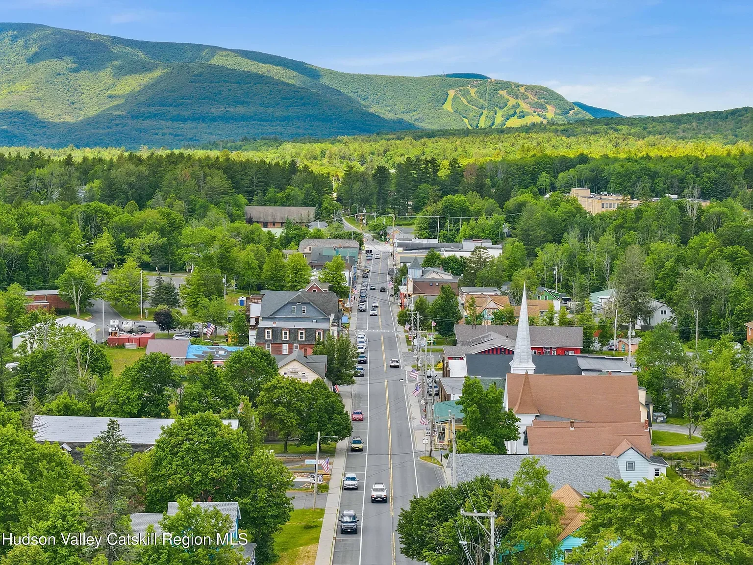 A small town with a main street running through the center, surrounded by lush green trees and hills in the background, under a blue sky.