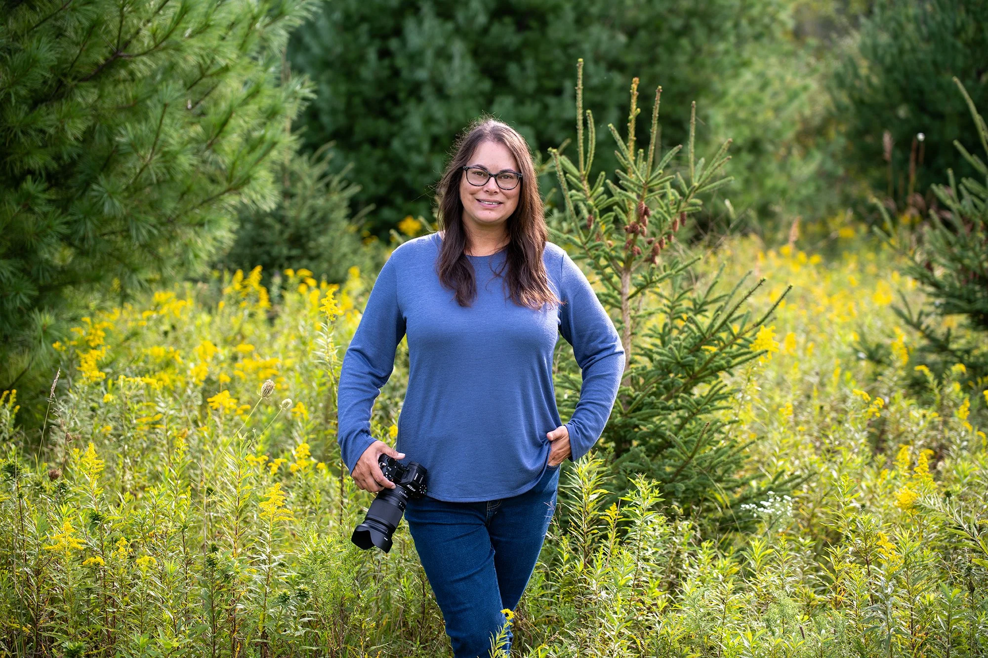A woman with glasses and long brown hair, wearing a blue long-sleeve shirt and jeans, stands outdoors in a green, wooded area with tall plants and yellow flowers, holding a camera in her right hand and smiling.