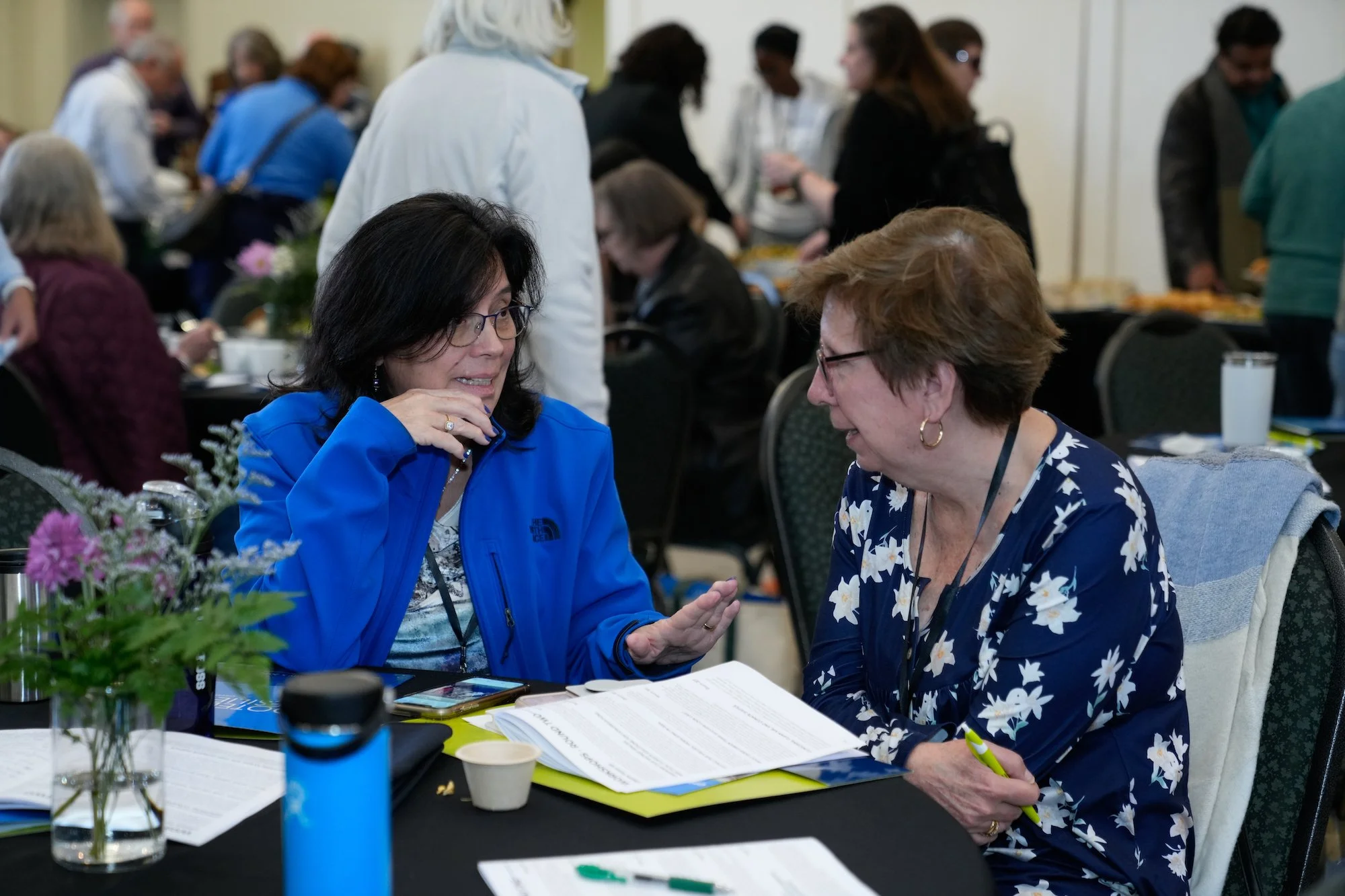 Two women with glasses and short hair are sitting at a table in conversation at a busy conference or event. They are surrounded by papers, notebooks, and drinks, with others standing and sitting in the background.