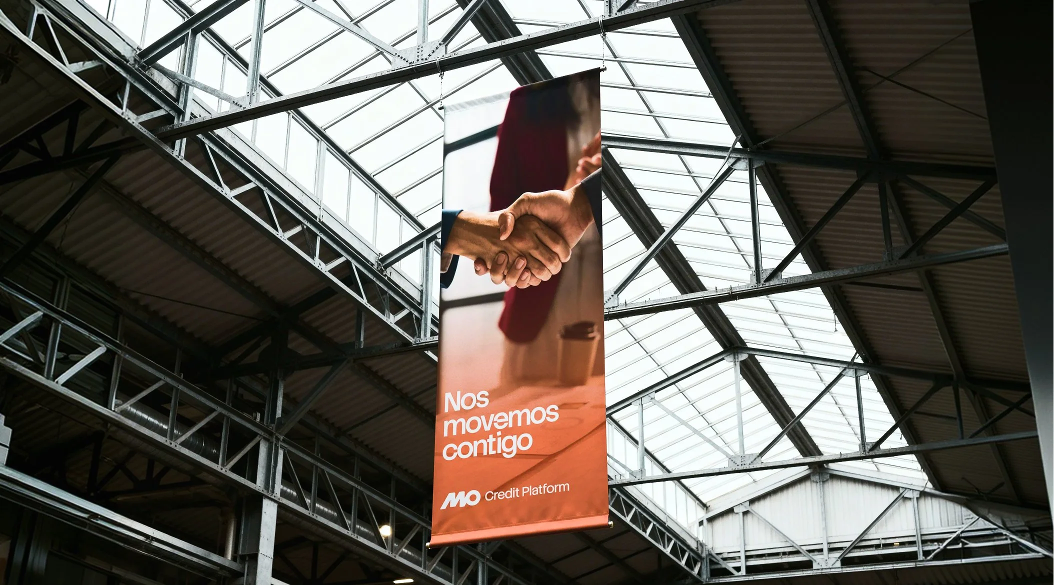 Puente metálico en un techo de cristal con una pancarta naranja que muestra la mano de dos personas dándose la mano y el texto "Nos movemos contigo".