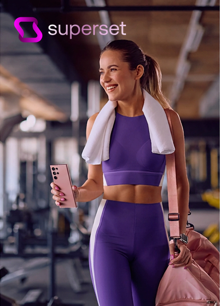 Mujer sonriente con ropa deportiva en un gimnasio, sosteniendo un teléfono móvil, con una toalla y mochila.
