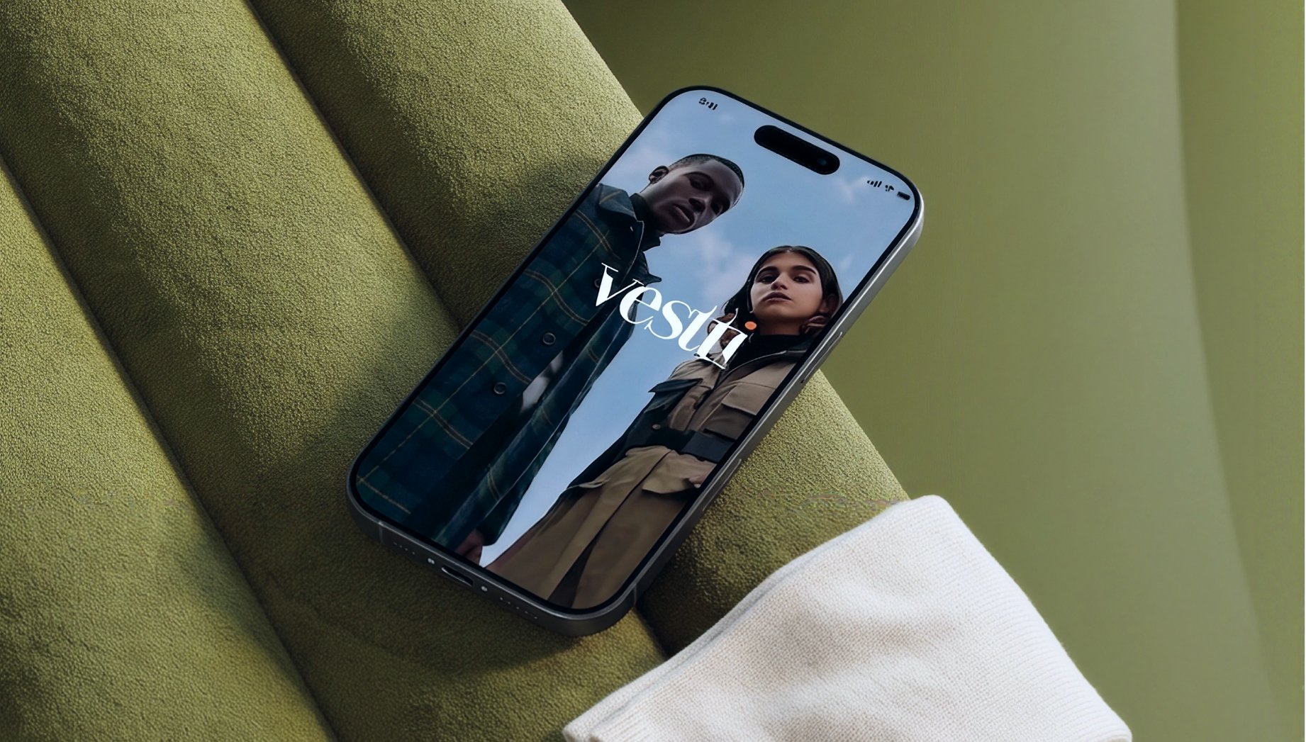Teléfono inteligente con una imagen en la pantalla de dos personas, un hombre y una mujer, vestidas con prendas de color oscuro y beige respectivamente. La pantalla muestra la palabra 'Vesti' y el teléfono está colocado sobre un sillón verde.