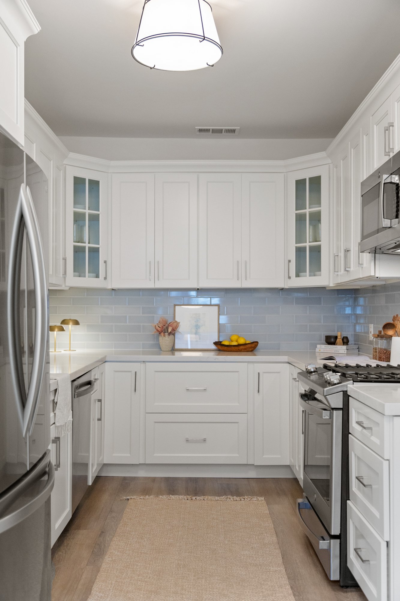 A modern white kitchen with white cabinets, stainless steel appliances, a beige counter, lemon bowl, pink flowers, and decorative items on the counter. Recessed lighting and crown molding are above.