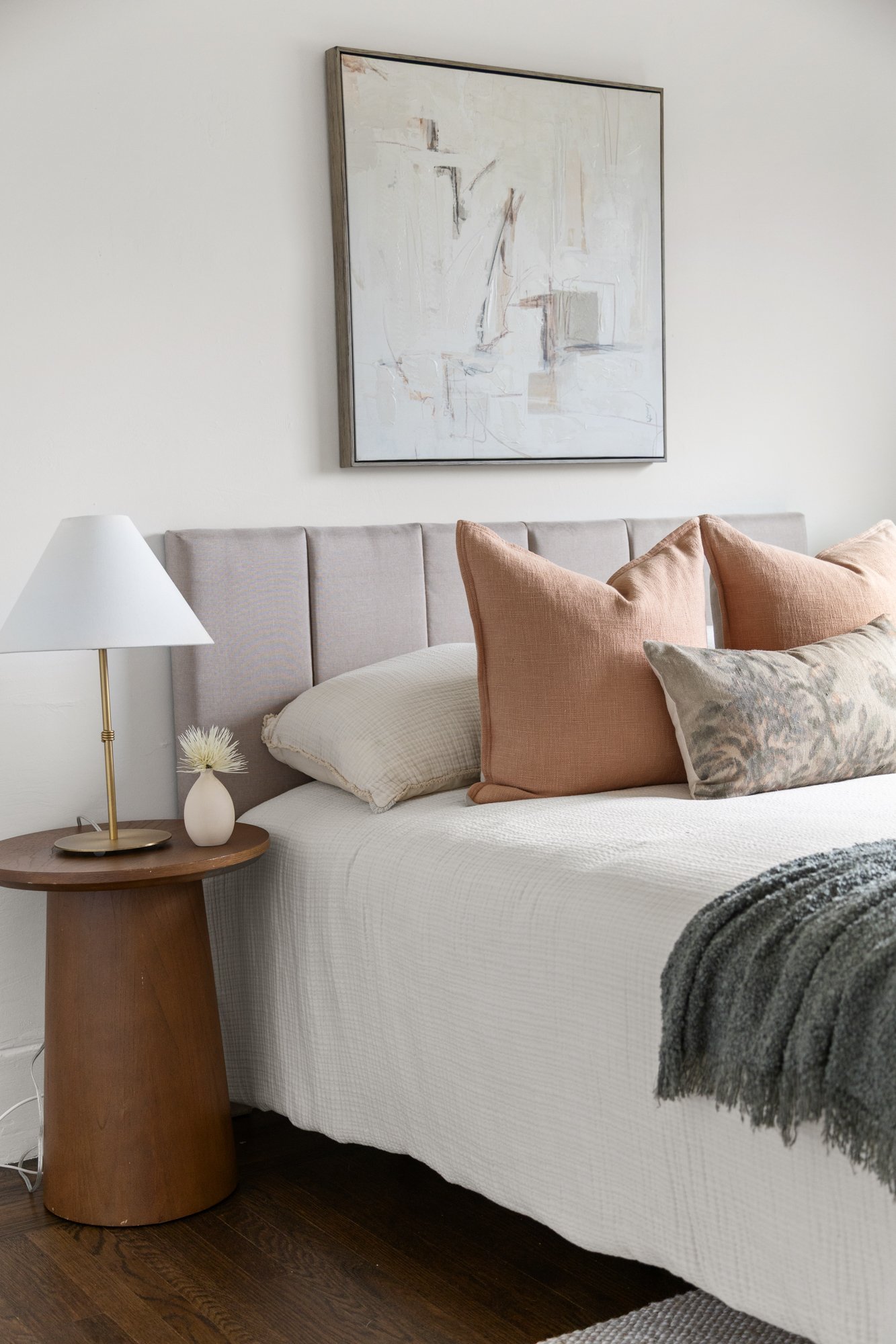 A bedroom with a white bed, beige and gray pillows, a gray blanket, a wooden nightstand with a table lamp and a small vase of flowers, a large abstract painting on the wall, and dark hardwood floors.