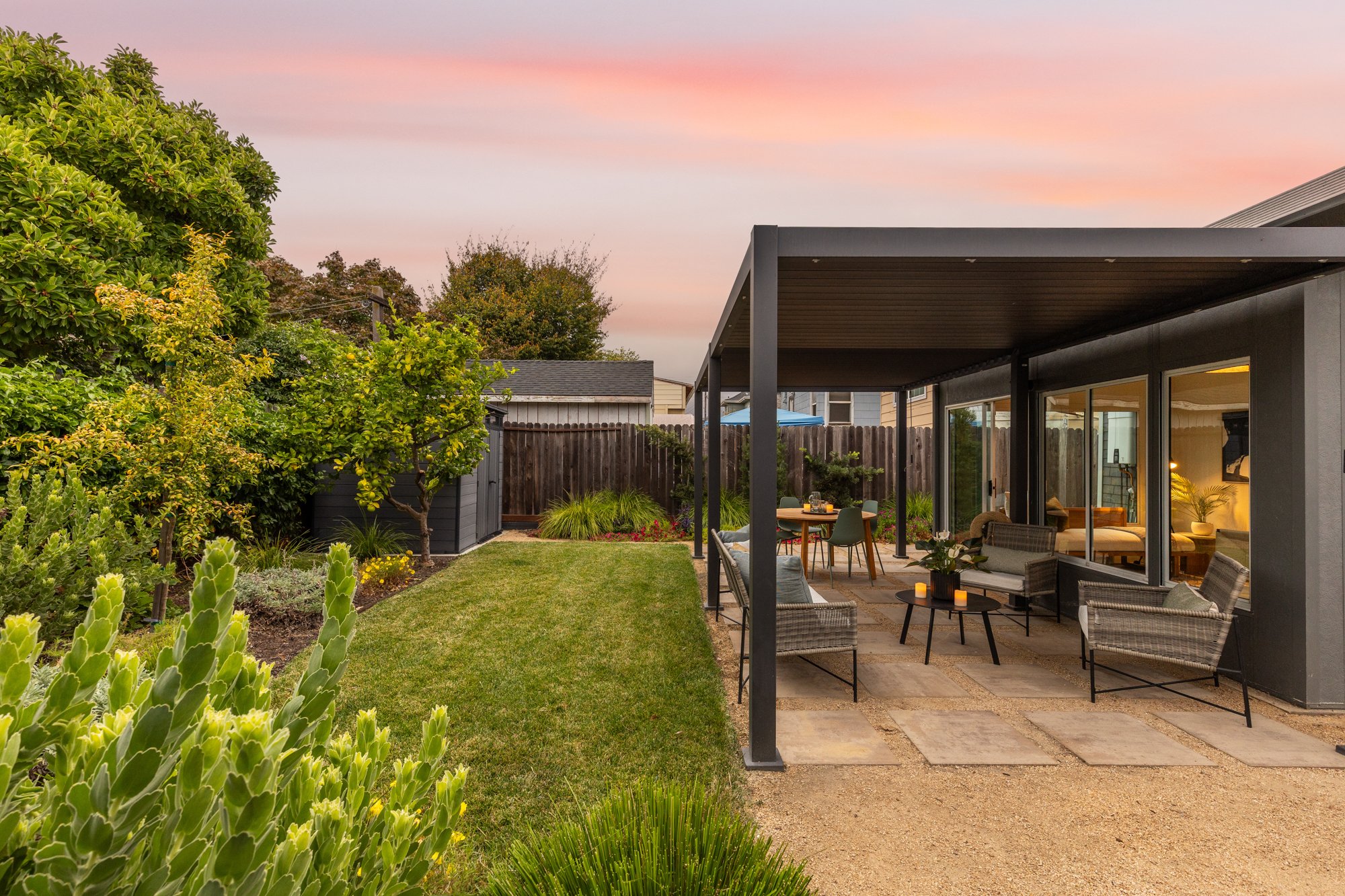 A backyard patio with outdoor seating, patio table, and candles, surrounded by lush green plants and trees, under a colorful pink and orange sunset sky.