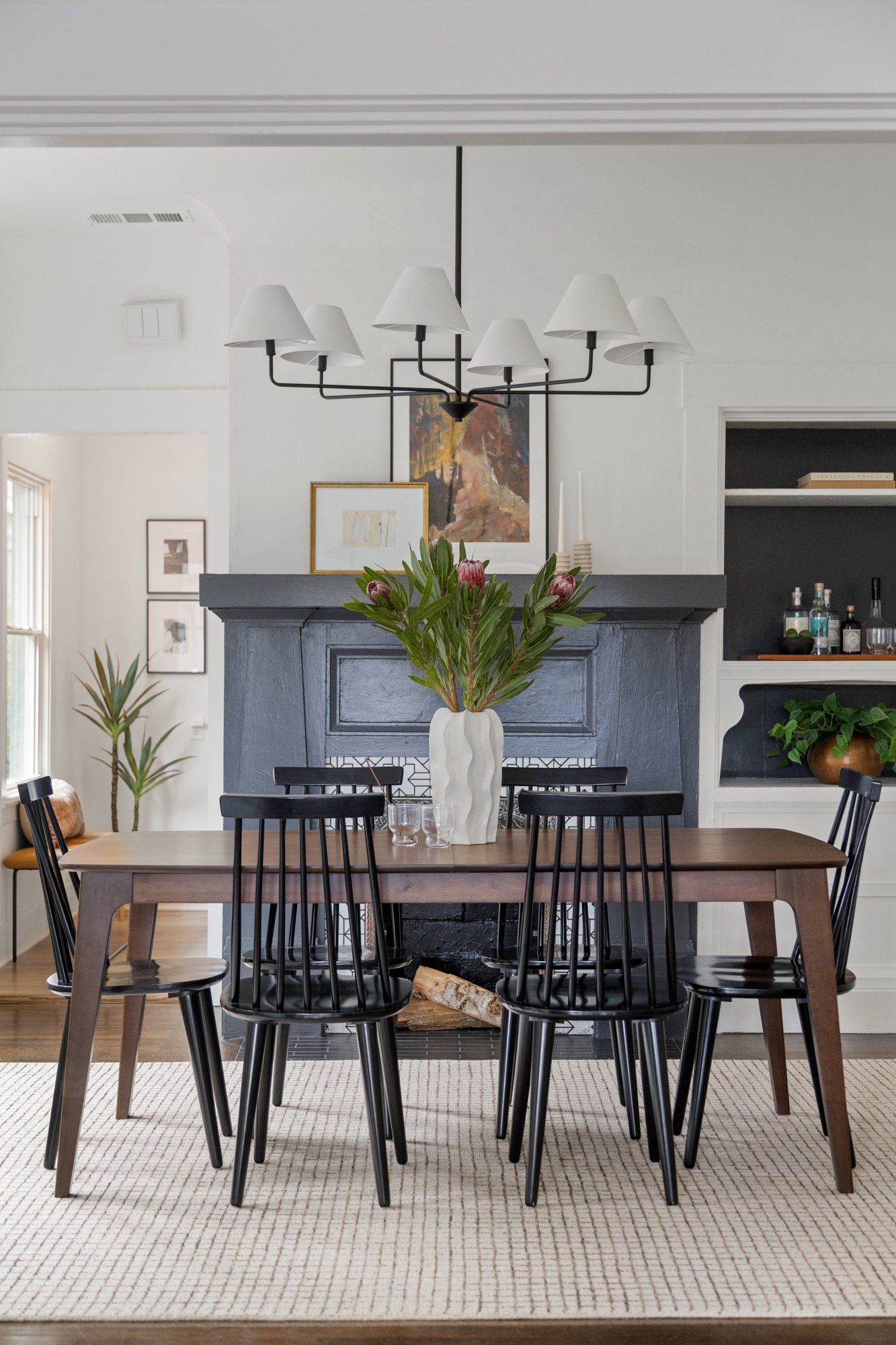 Dining room with a wooden table, black chairs, a white ceramic vase with pink flowers, a fireplace, artwork on the wall, and a chandelier with nine lampshades.