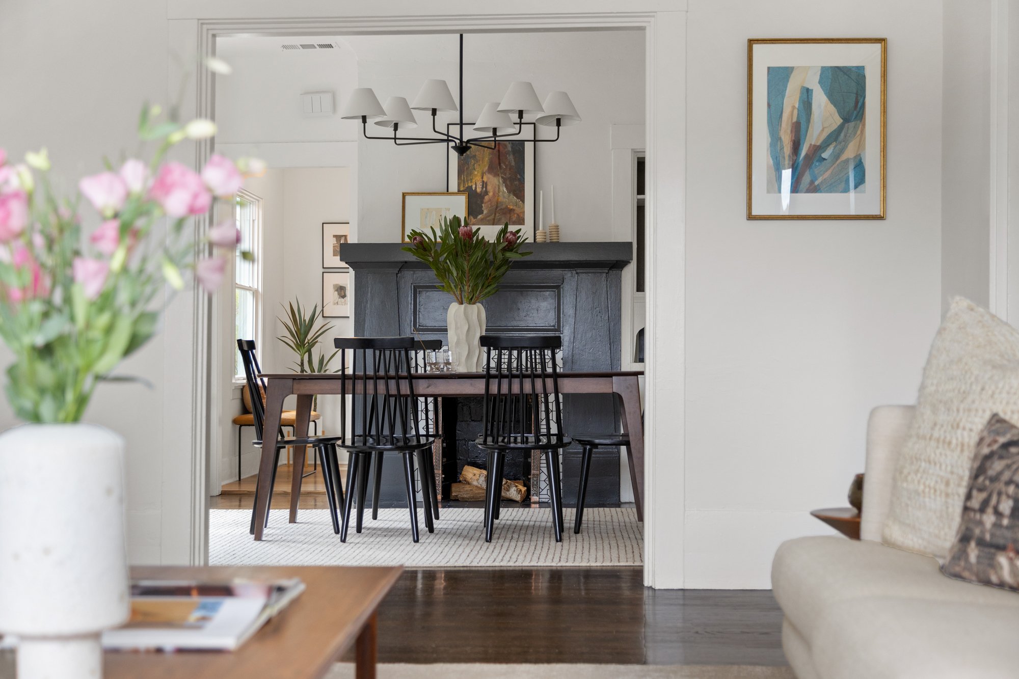 Living room with a dining area & fireplace, decorated with framed artwork, a chandelier, and a vase of pink flowers in foreground.