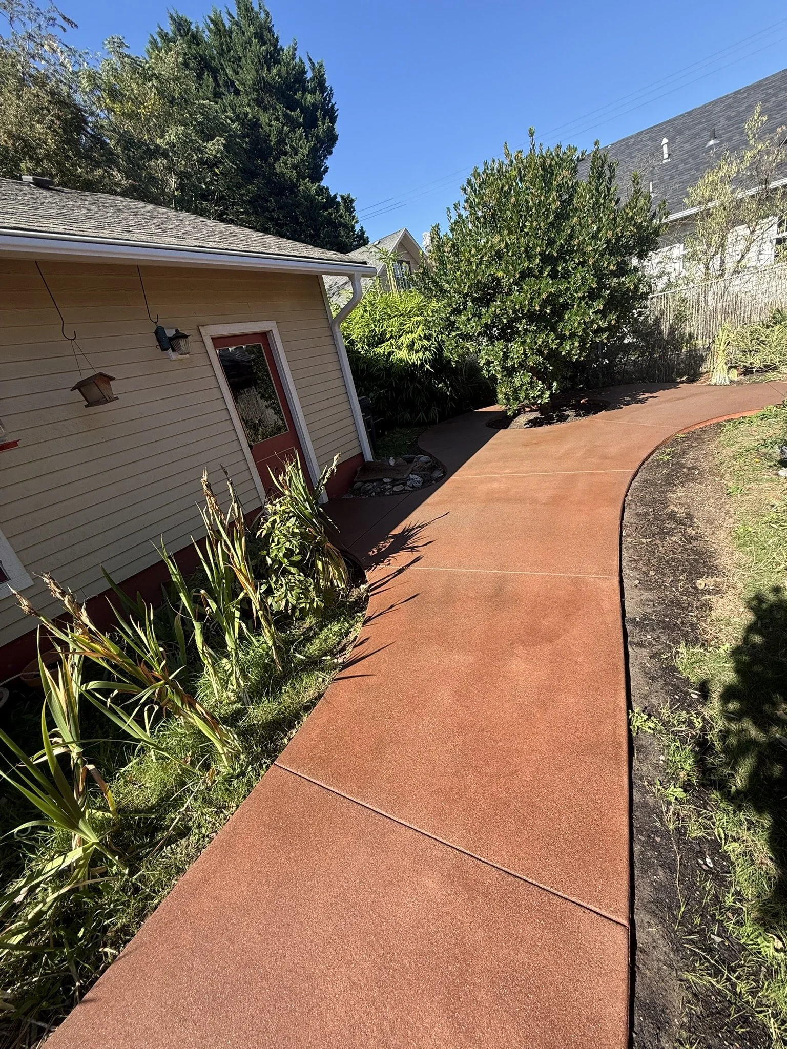 Reddish concrete walkway curving through a garden beside a small building.