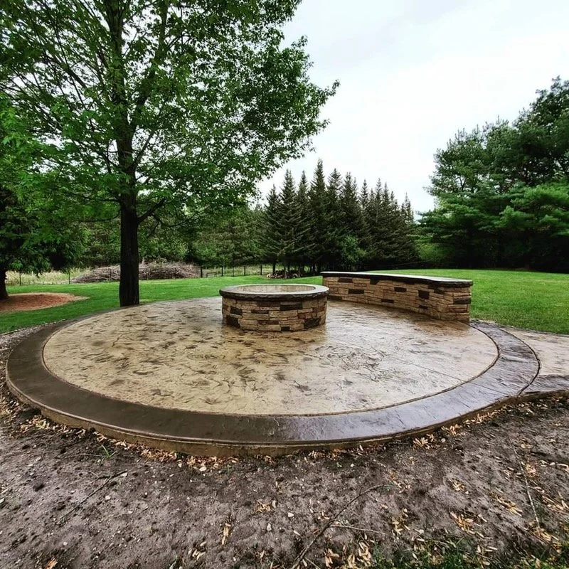 Circular concrete patio with a contrasting dark border and a stone fire pit in a landscaped yard.