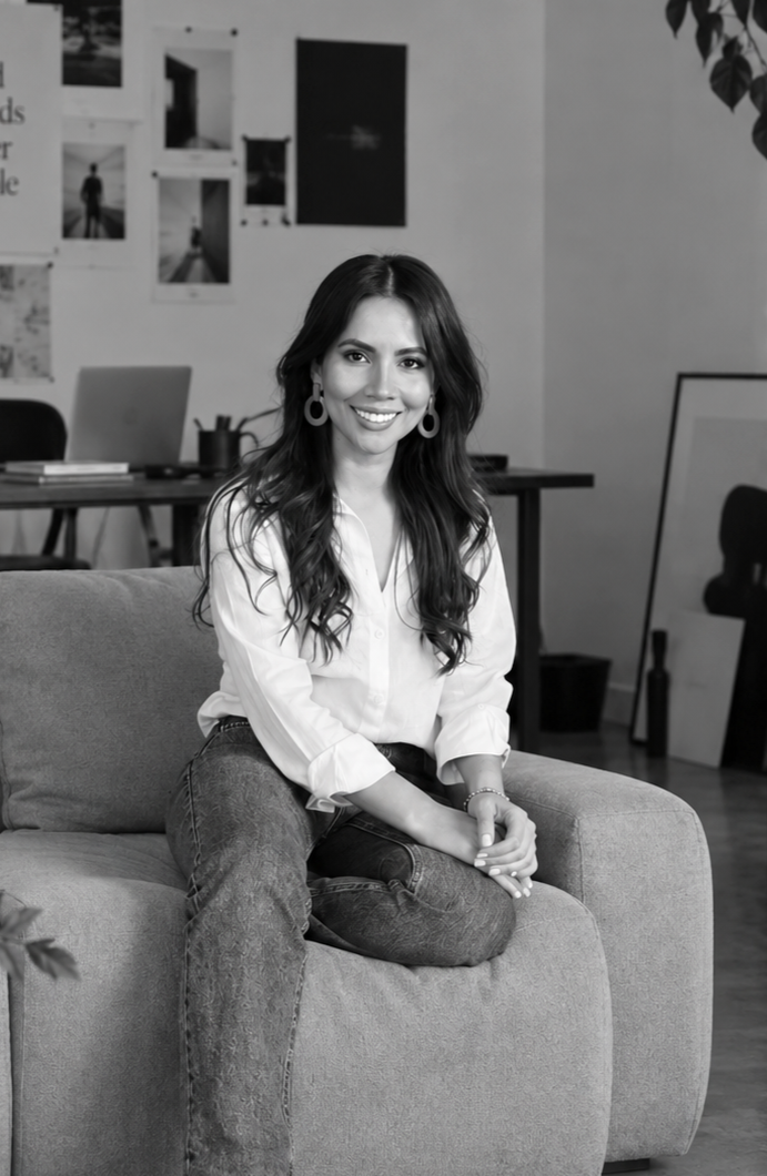 Mujer sonriente con cabello rizado, usando camisa blanca y jeans, sentada en un sofá.