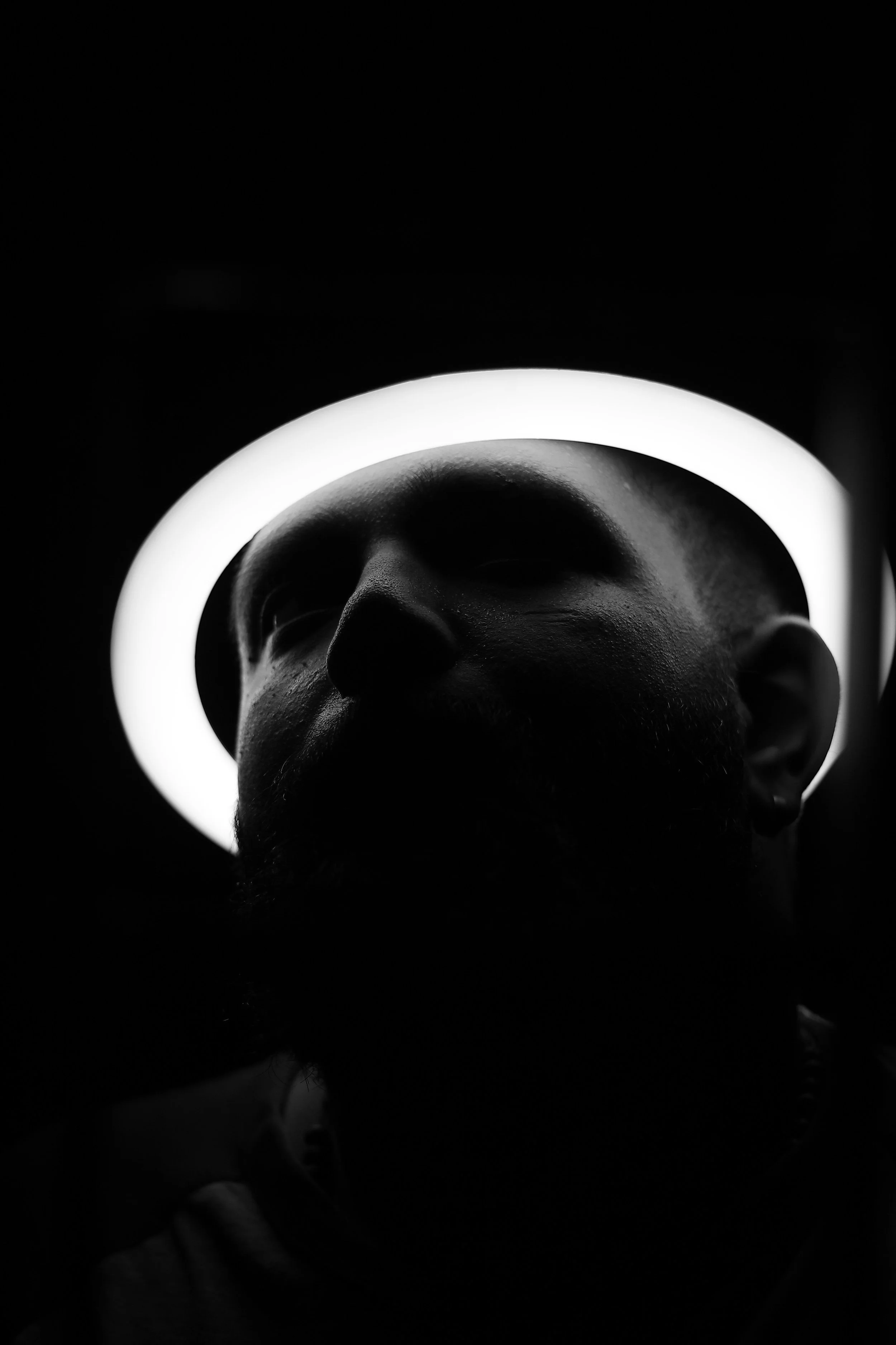 Una figura de una cabeza humana con barba, vista desde abajo, rodeada por un halo de luz blanca en forma de círculo, en fondo oscuro.