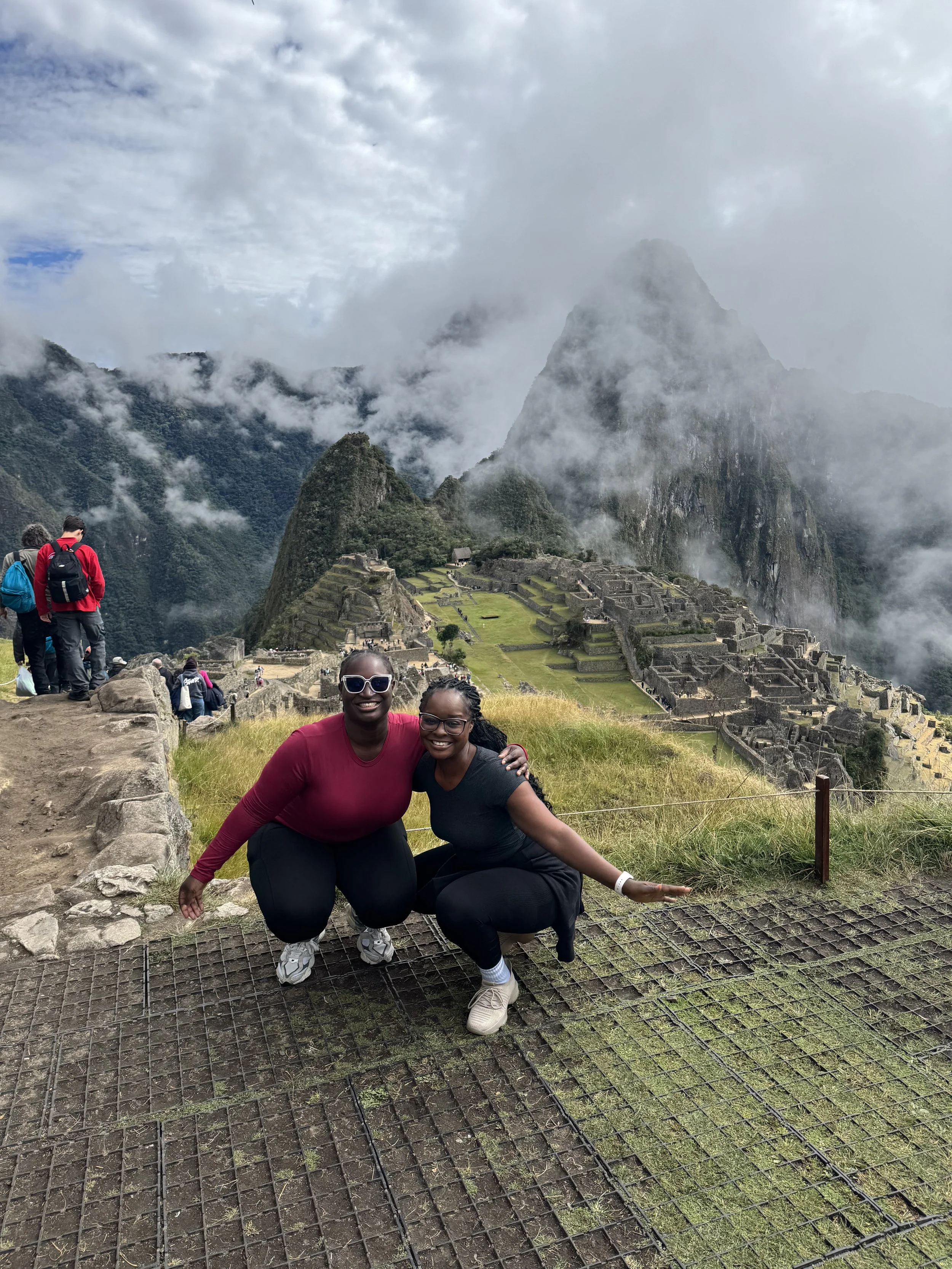 Two women posing and smiling in front of Machu Picchu, with tourists and the ruins visible in the background, surrounded by mountains and clouds.