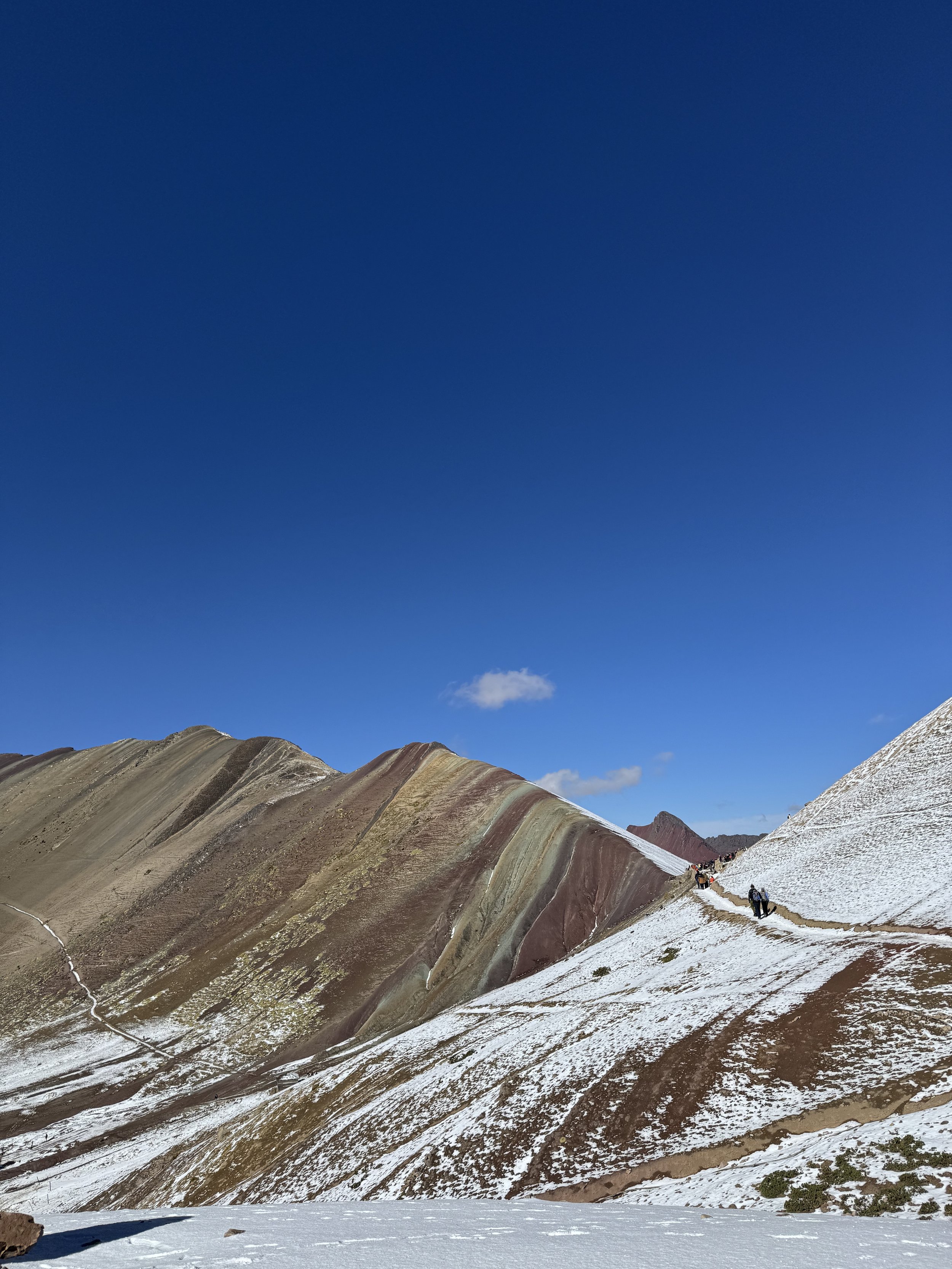 Colorful striped mountains with snow on some slopes, blue sky with a few clouds, and a trail with hikers walking along the right side of the mountain range.