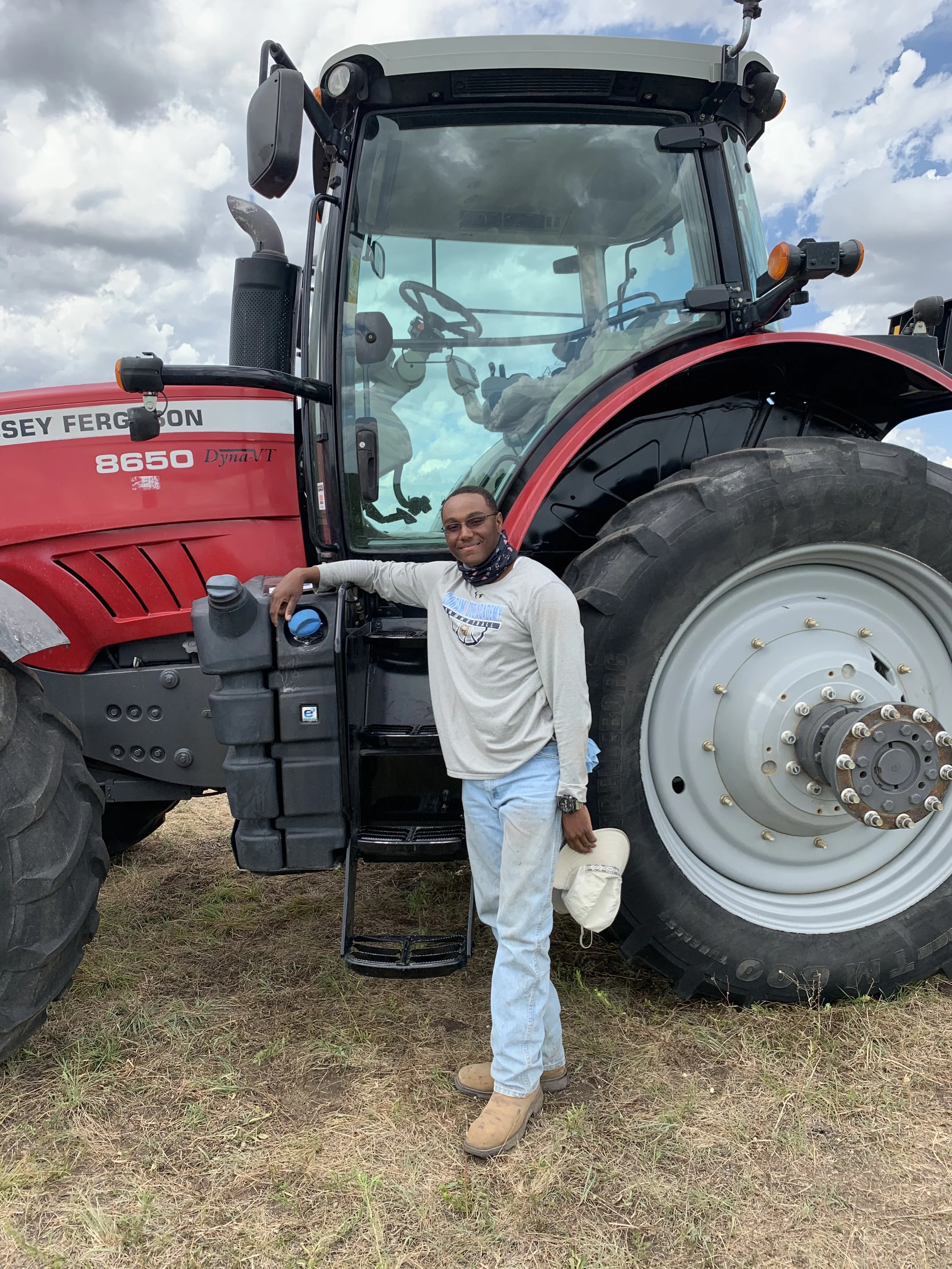 A man standing next to a large red Massey Ferguson tractor, smiling, with his hand resting on the tractor's control panel. The tractor has large tires and is outdoors on a grassy area under a partly cloudy sky.