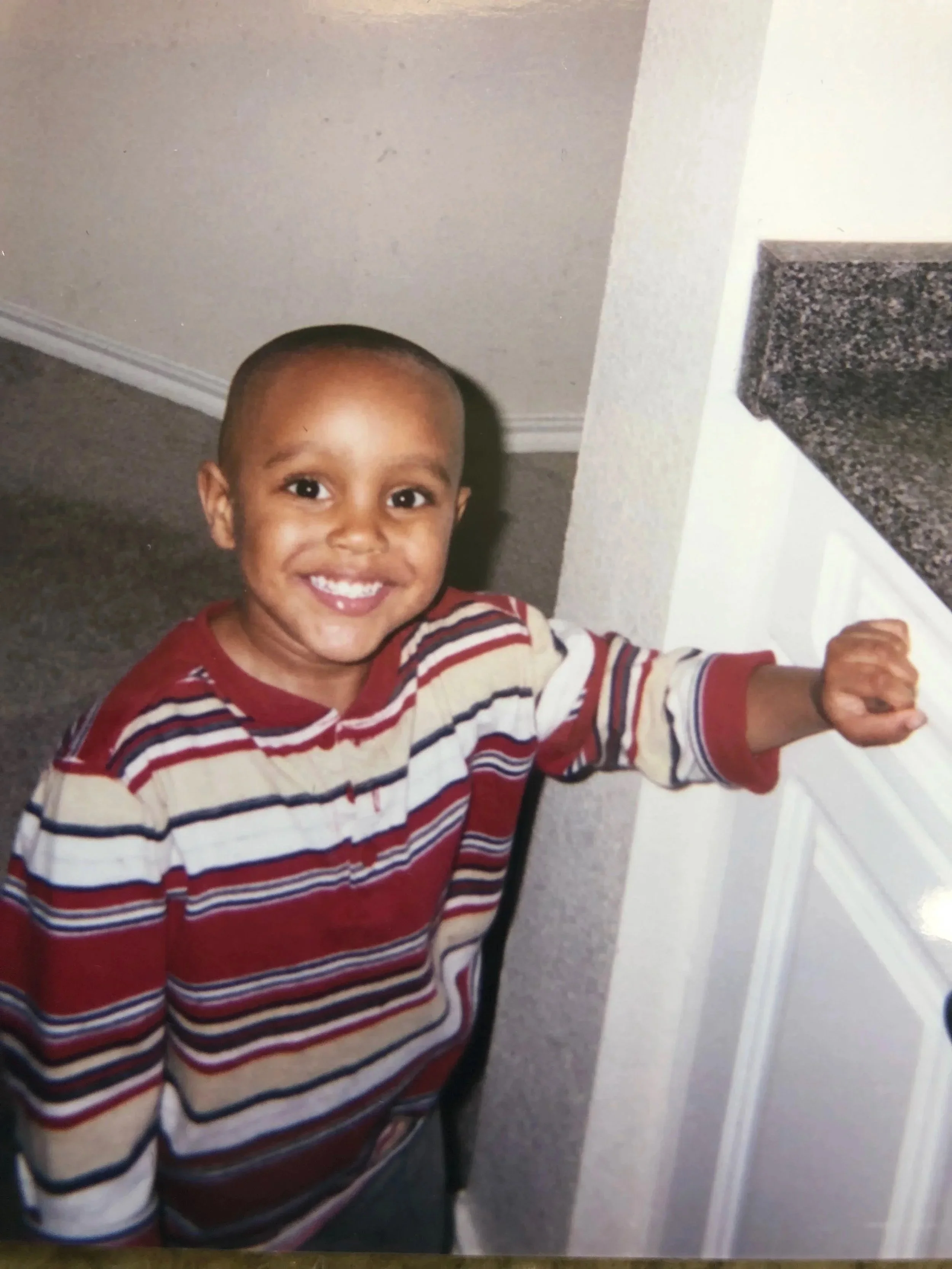 A smiling young boy with a shaved head, wearing a striped red, white, and beige sweater, reaching towards a white cabinet in a room with gray carpet.