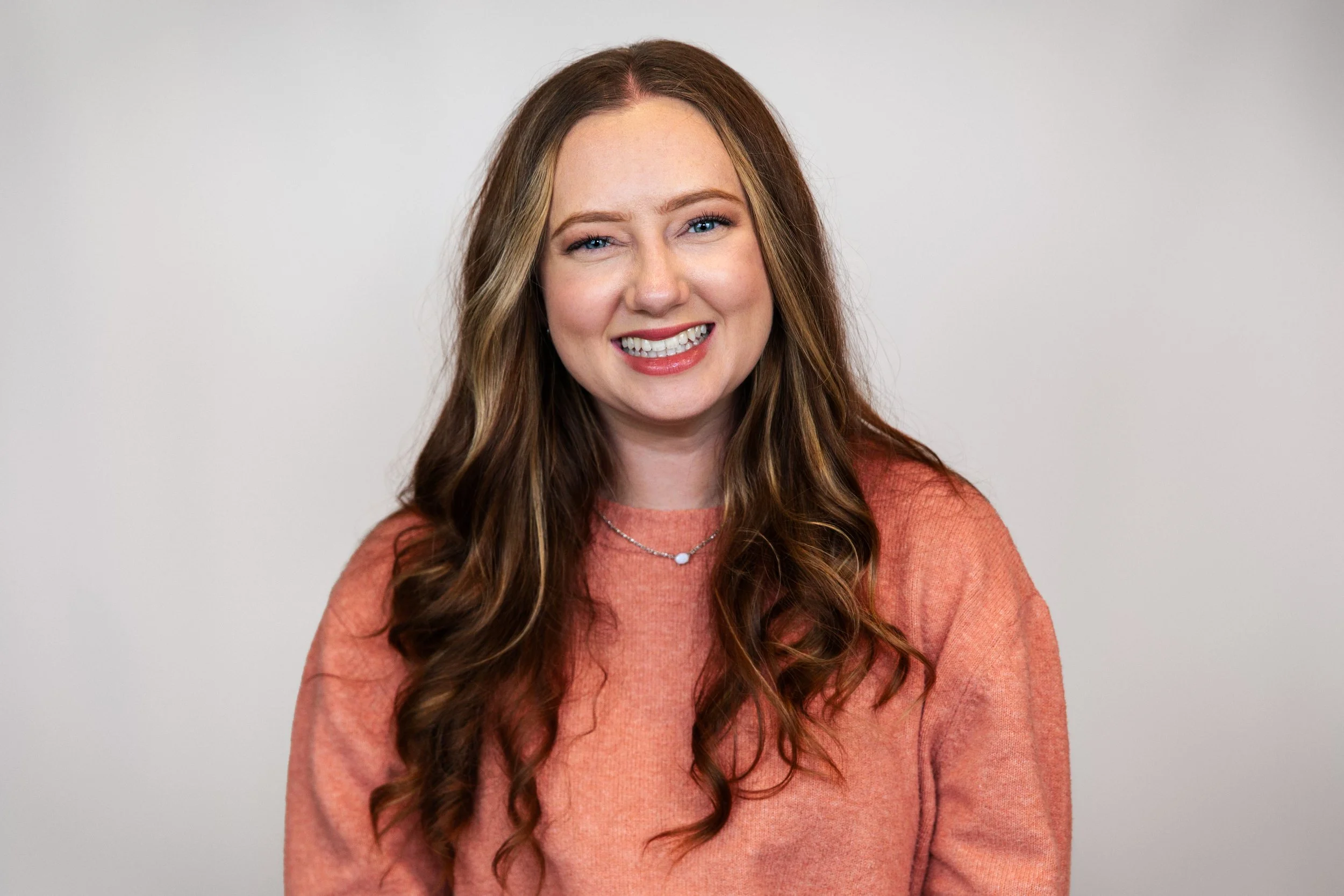 A smiling woman with long, wavy, auburn hair, wearing a peach-colored sweater and a delicate necklace, standing against a plain off-white background.