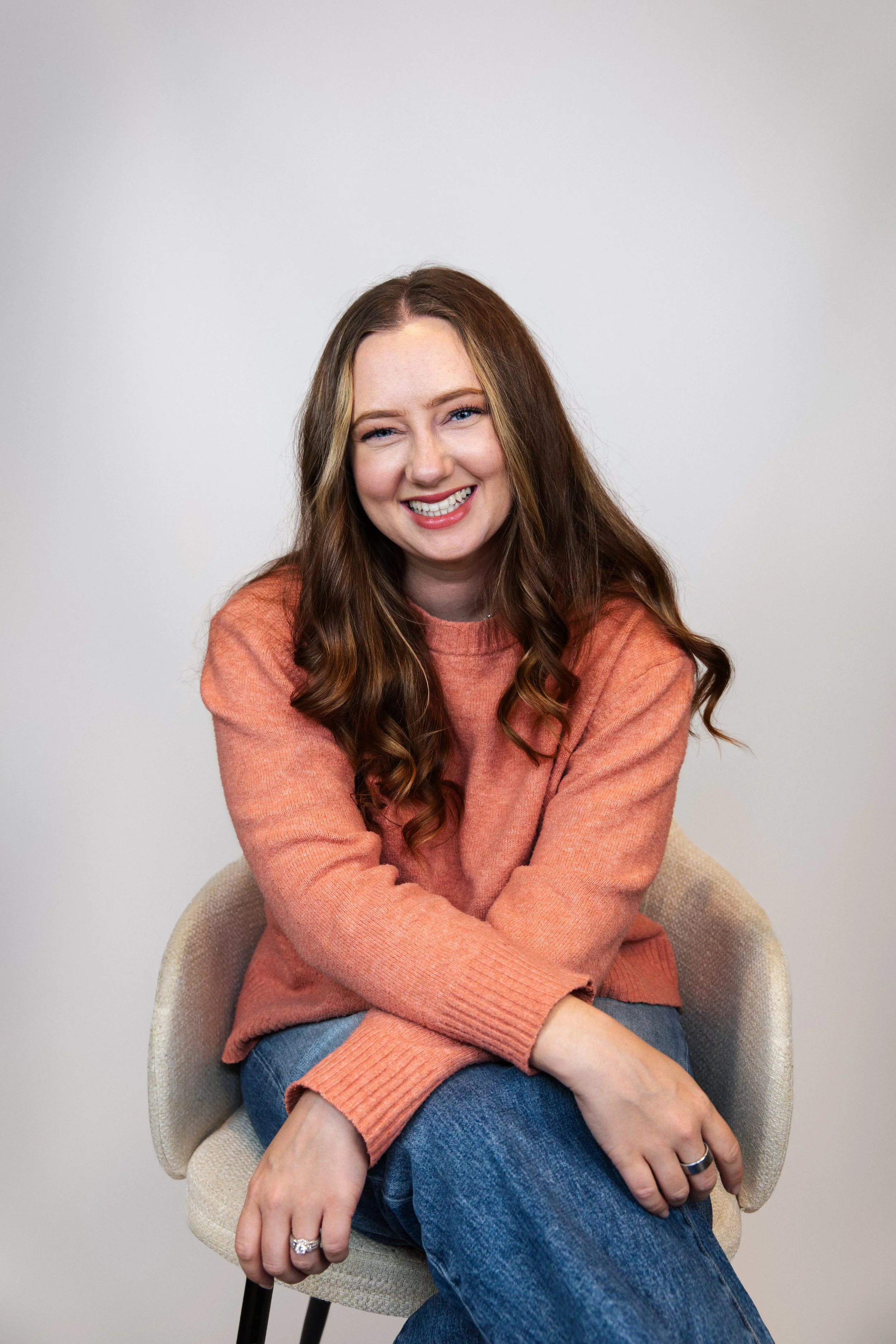 A young woman with long wavy brown hair, wearing an orange sweater and blue jeans, sitting on a beige chair in front of a plain light gray background, smiling and looking at the camera.