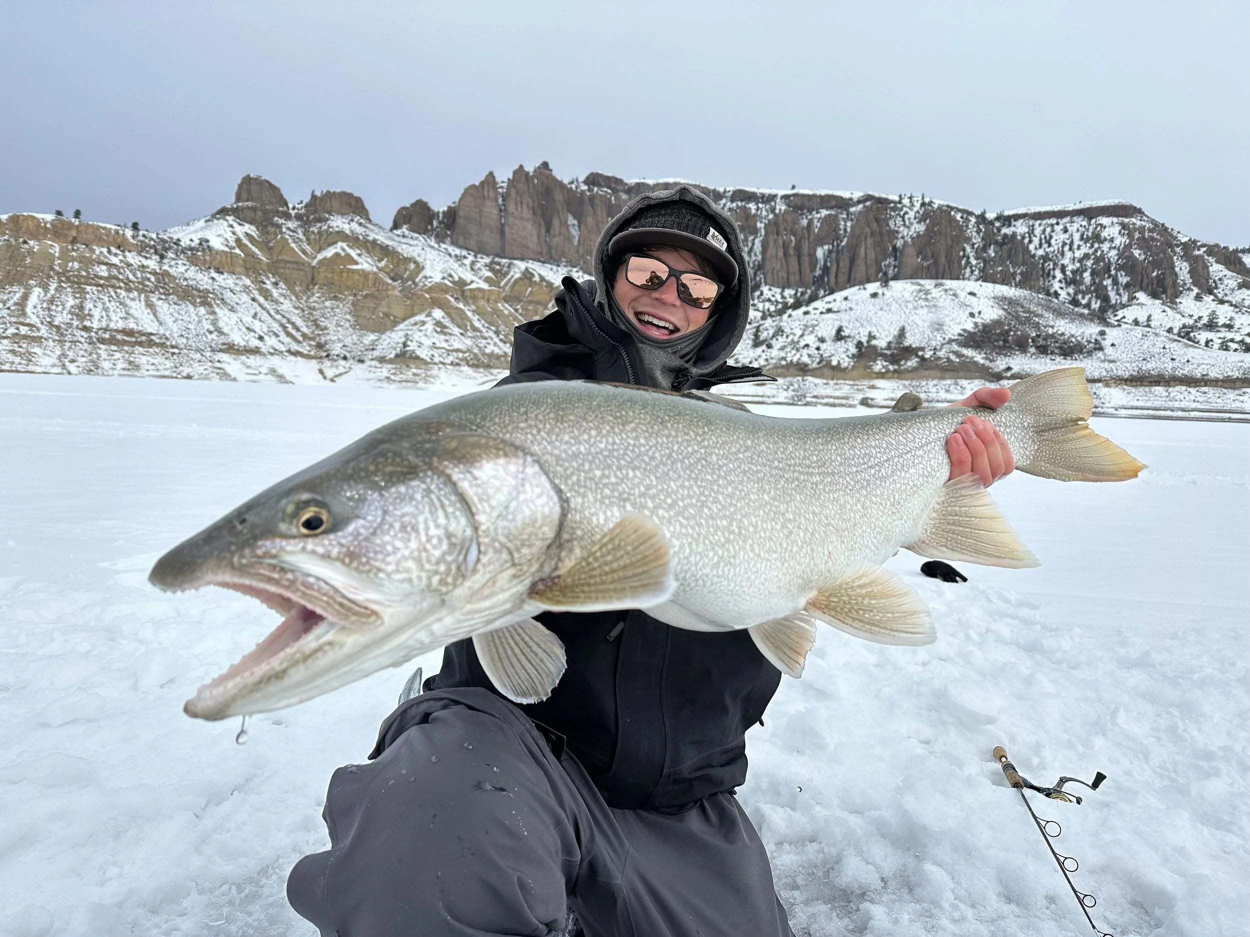 Person holding a large fish on a snowy landscape, with snow-covered mountains in the background, wearing winter gear including sunglasses and a jacket.