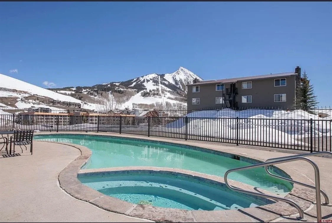 Outdoor heated swimming pool with stairs, surrounded by a fence, with a backdrop of snowy mountains and a gray apartment building under a blue sky.