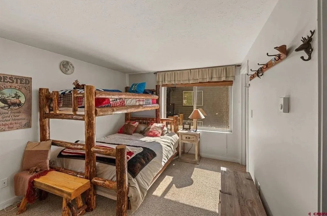 A rustic-style bedroom with a log bunk bed, a small nightstand with a lamp, a window with beige curtains, and wall hooks shaped like a reindeer and a stag head.