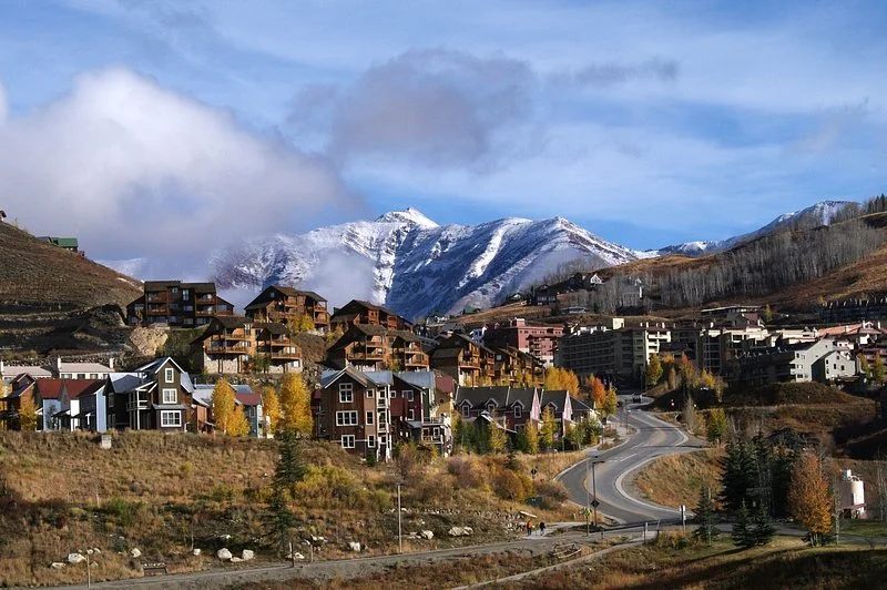 Hillside town with colorful houses and winding road, mountain range with snow-capped peaks in the background, some trees with fall foliage.