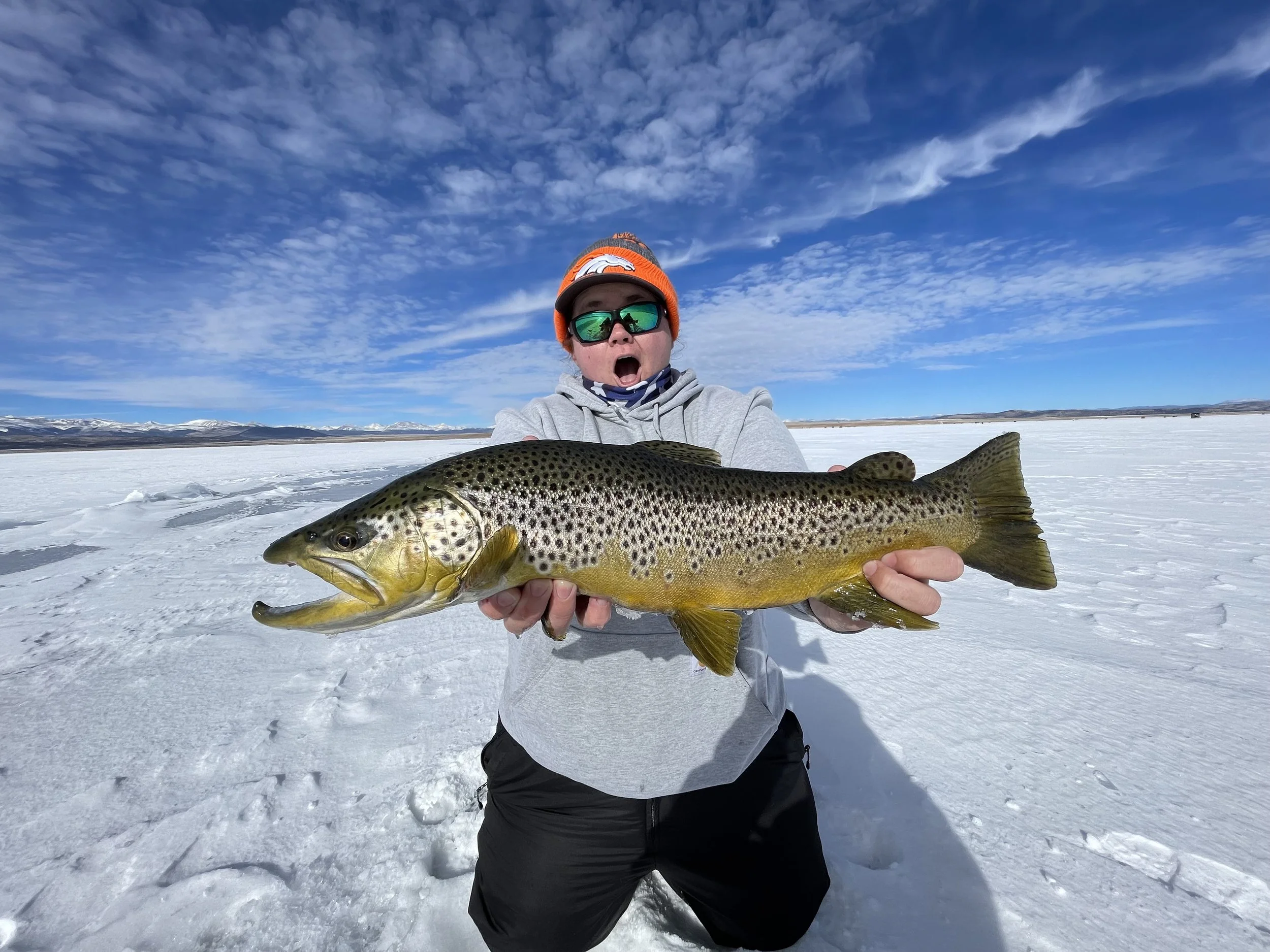 Person wearing sunglasses and an orange beanie holding a fish on a snowy landscape with mountains and a blue sky with clouds in the background.