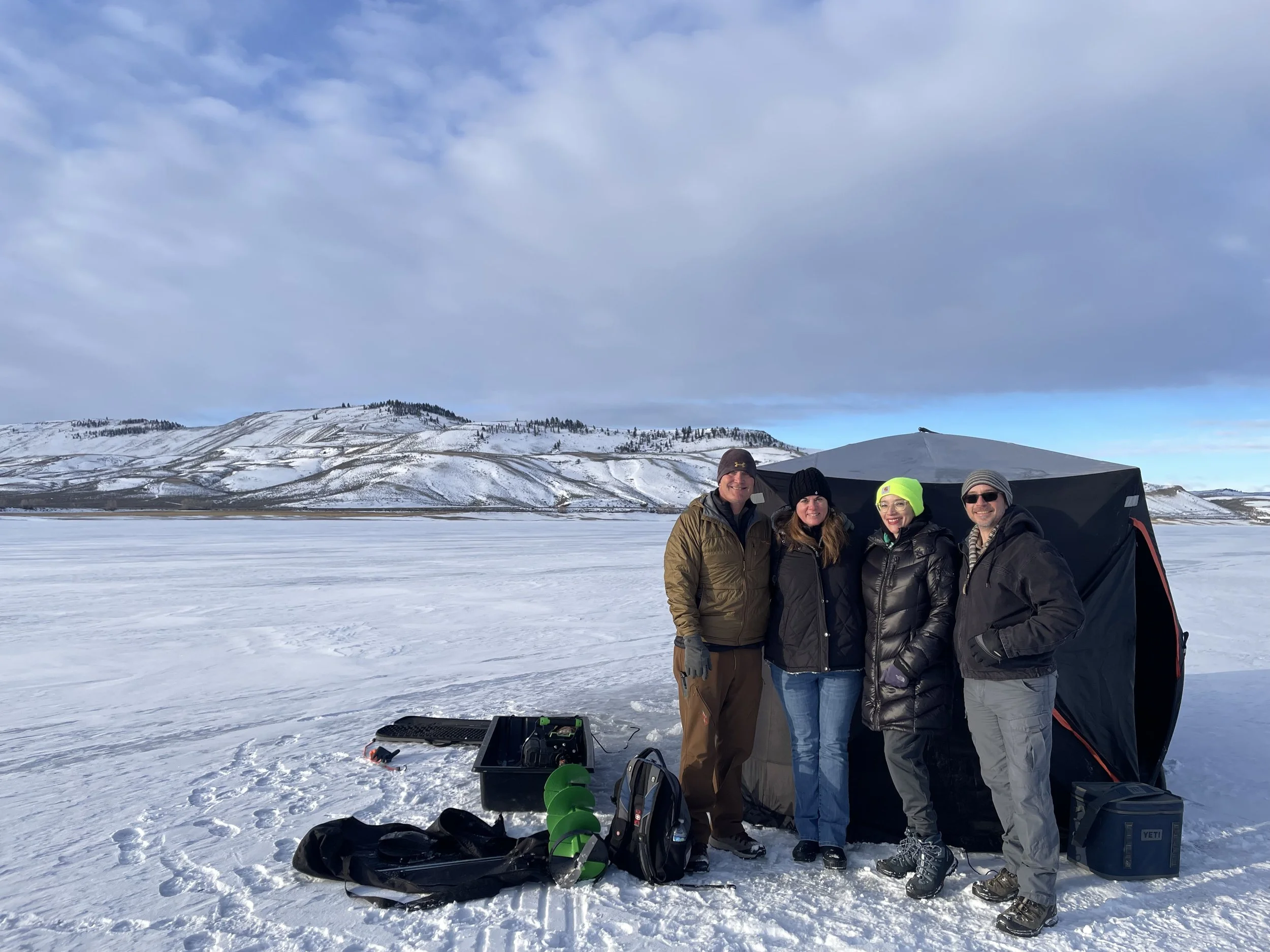 A family of four people standing outdoors on a snowy landscape with mountains and cloudy sky in the background, smiling, wearing winter clothing. Equipment and supplies for outdoor activity are visible on the ground.