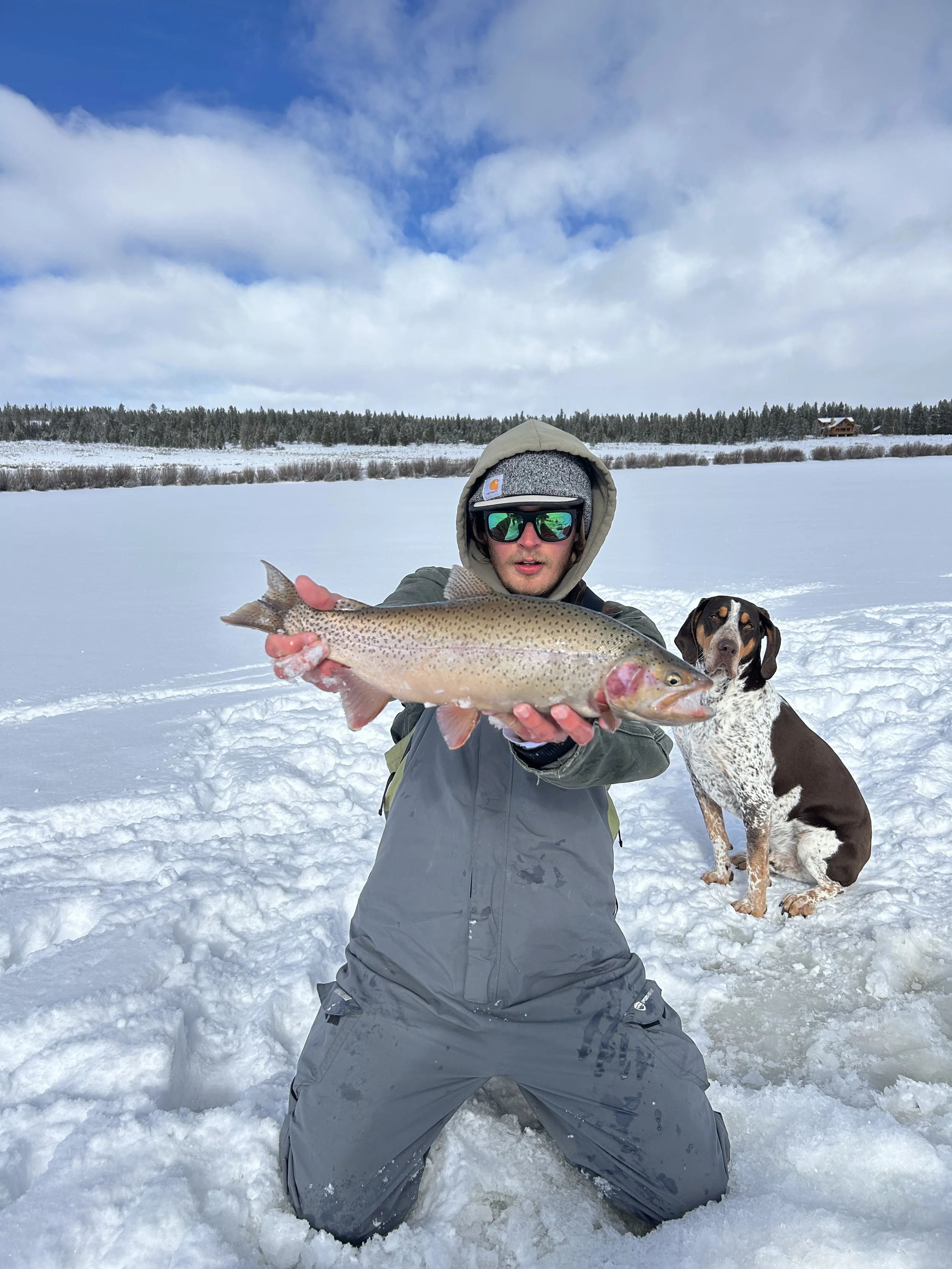 A person kneeling in snow holding a large fish, with a dog sitting nearby on snow-covered ground, and a winter landscape with trees and a house in the background.