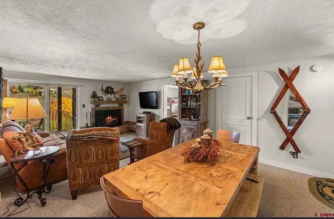 Living room with a wooden dining table, leather chairs, a sofa, a fireplace, a TV, sliding glass doors leading to a balcony, and mountain view outside.