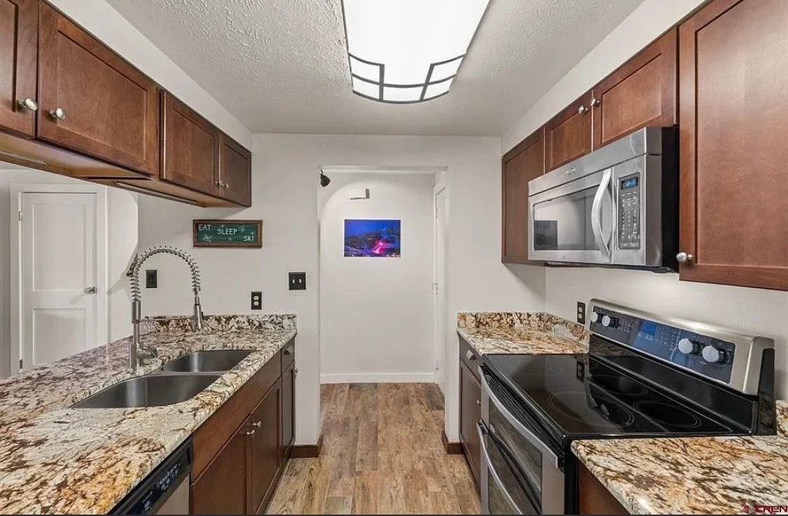 Kitchen with brown wooden cabinets, granite countertops, a double sink, a microwave, an oven, and a wooden floor.
