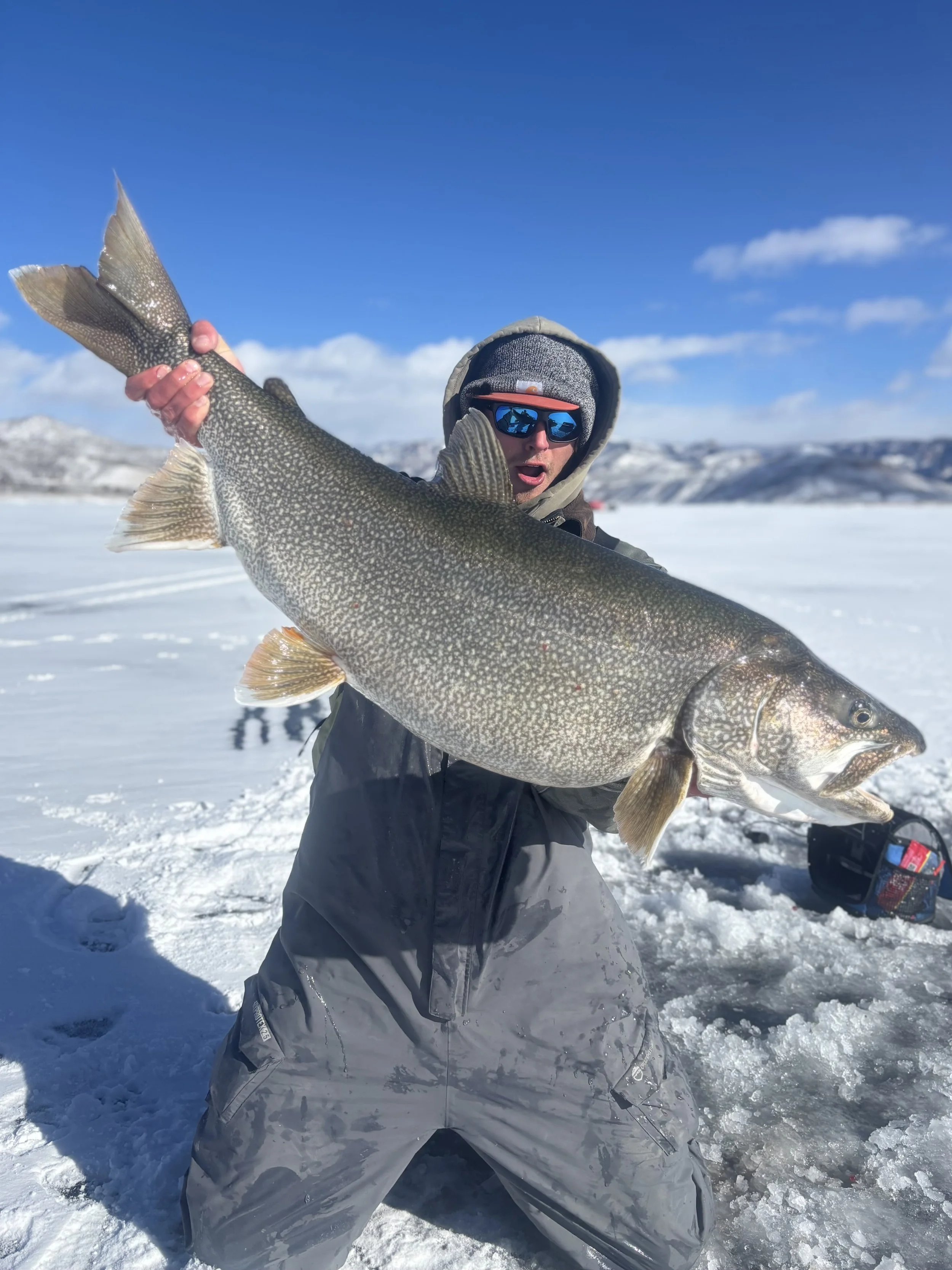 An angler wearing winter gear holds a large, trophy lake trout on Blue Mesa Reservoir, while ice-fishing in the Gunnison valley under snow-capped peaks.