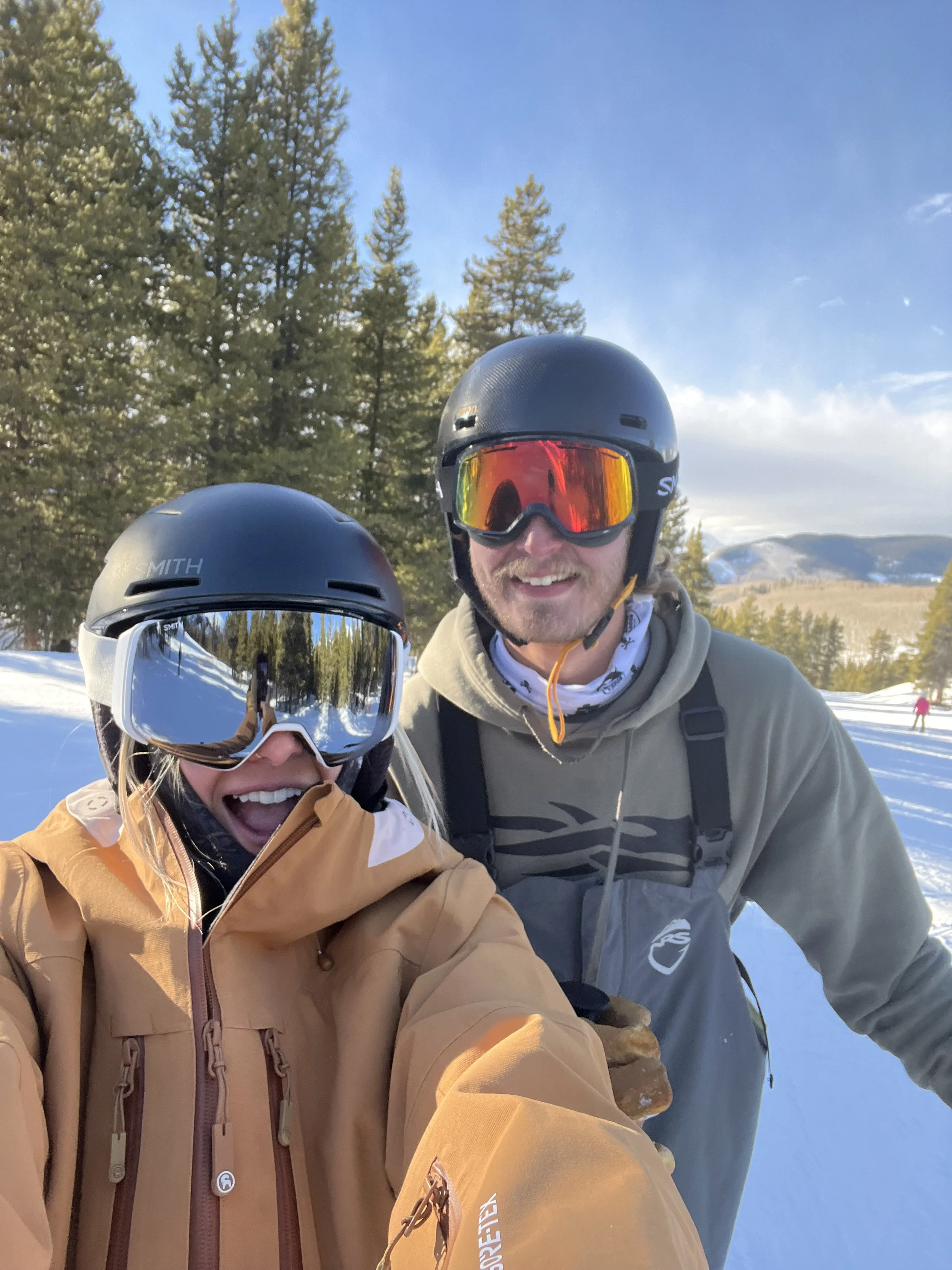 Two people wearing ski helmets and goggles smiling outdoors in a snowy area with trees and mountains in the background.