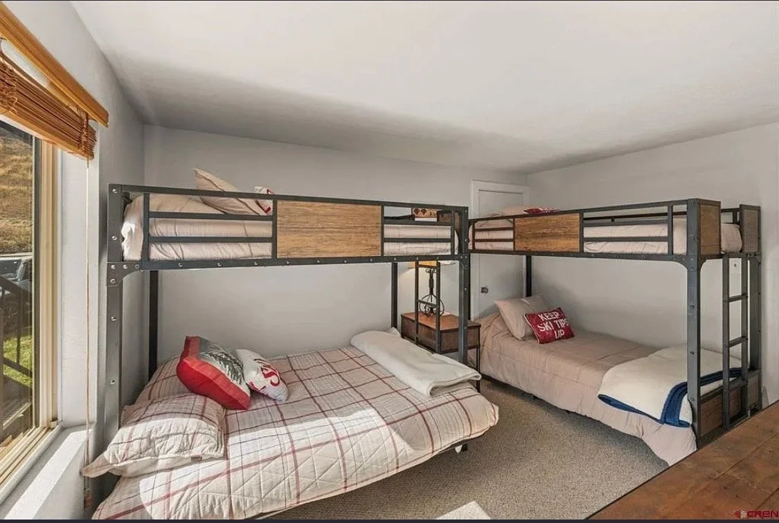 Bedroom with two sets of metal loft beds, each with twin mattresses and bedding. The lower beds are made with decorative pillows, and there is a wooden side table with a lamp between the beds. A window with wooden blinds is on the left, and the room has white walls and a beige carpet.