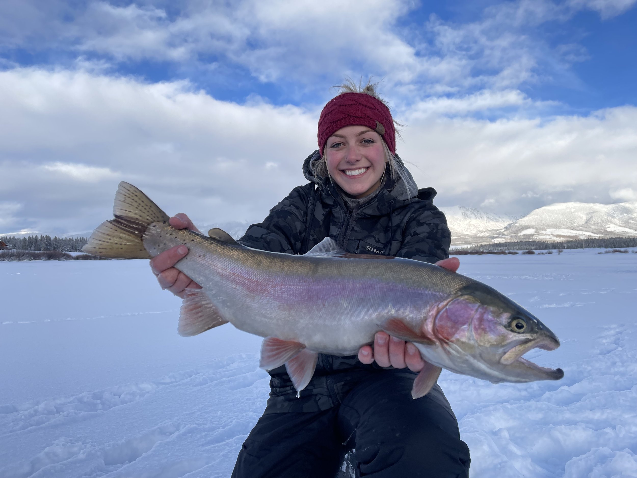 A young woman wearing a maroon knit hat and a black winter jacket smiles while holding a large fish against a snowy landscape with mountains and a partly cloudy sky in the background.