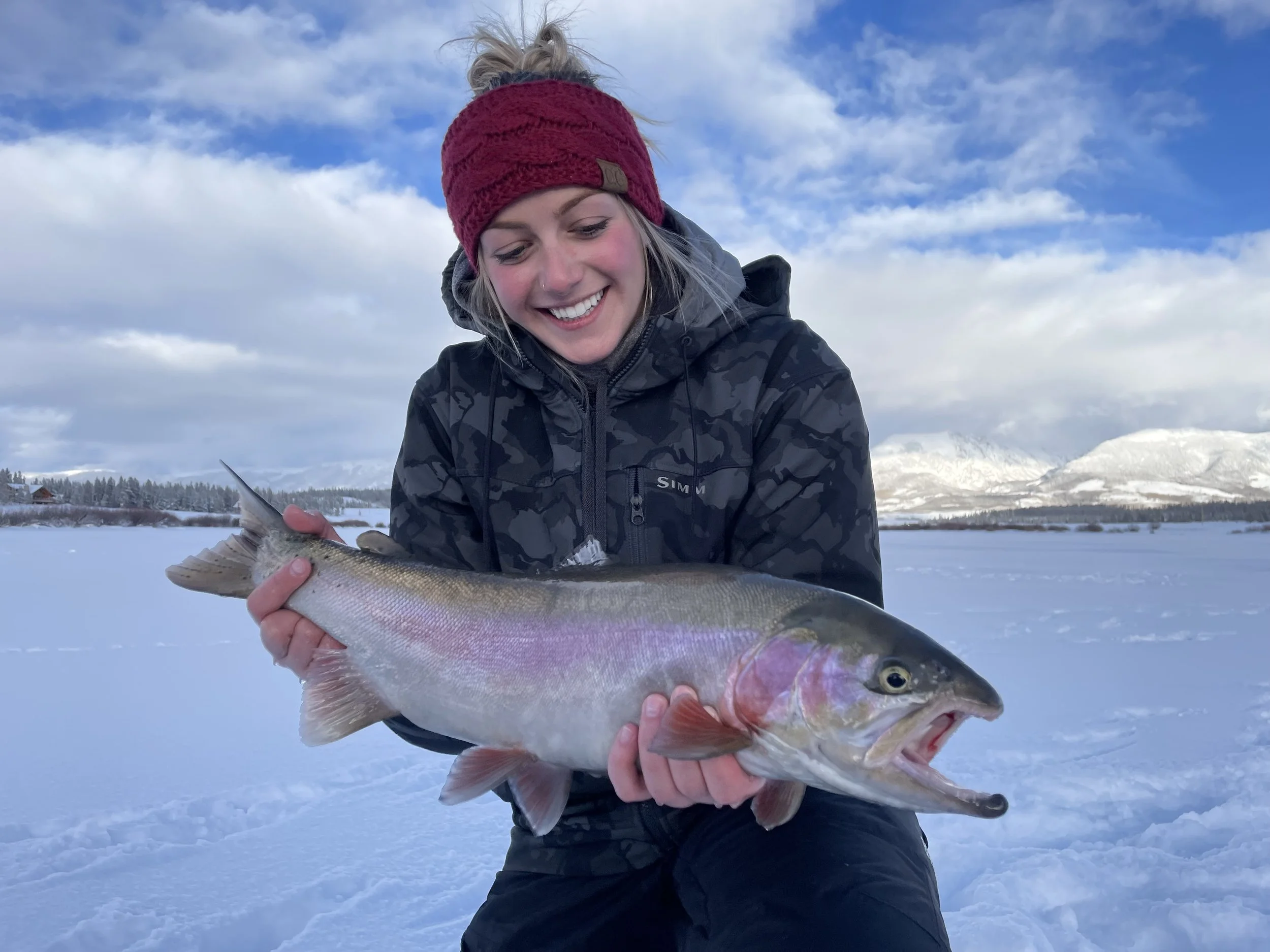 A woman in a red headband and black camouflage jacket smiling while holding a large fish in a snowy outdoor landscape with mountains and trees in the background.