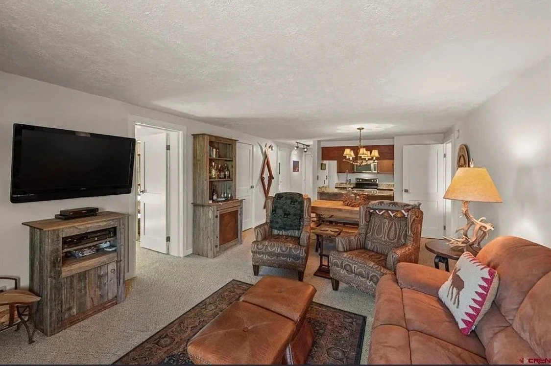 Living room with a mounted flat-screen TV, wooden furniture, patterned armchairs, a leather ottoman, and a sofa with a decorative pillow, leading to a kitchen area with a dining table and chandelier.