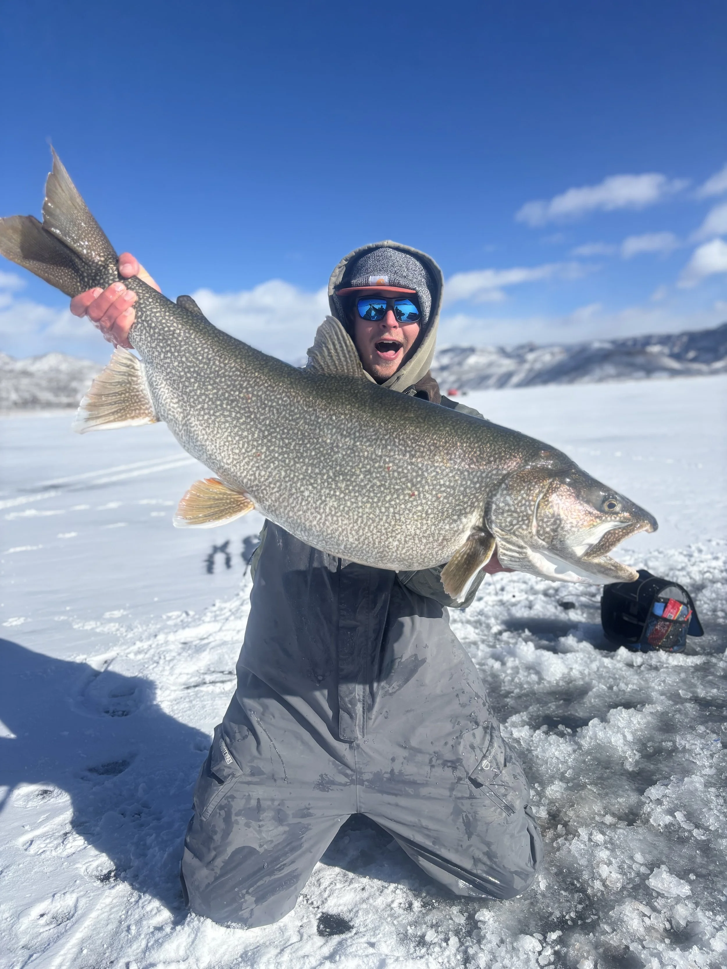 A person in winter clothing holding a large fish on a snowy landscape with mountains in the background.