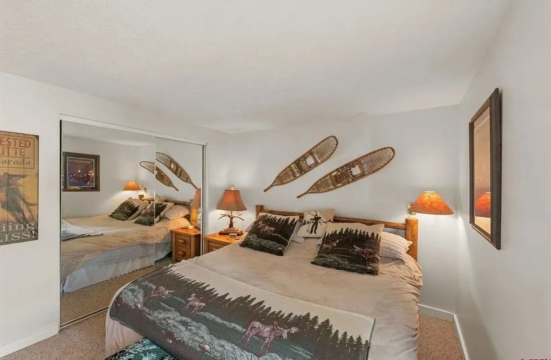 A cozy bedroom with a bed, wooden nightstands with lamps, wall decorations, and a mirror closet door reflecting the bed.