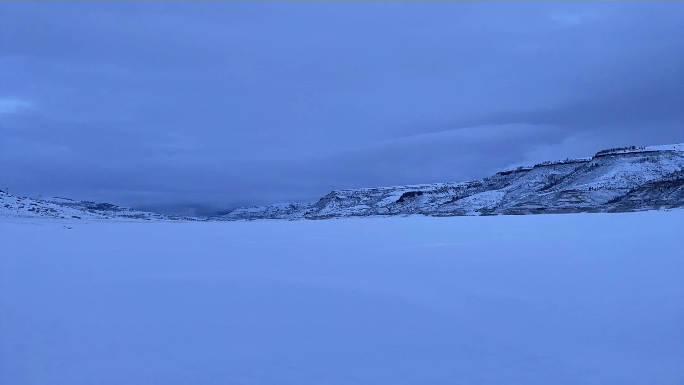 Cold winter landscape with snow-covered ground and mountains under a partly cloudy blue sky.