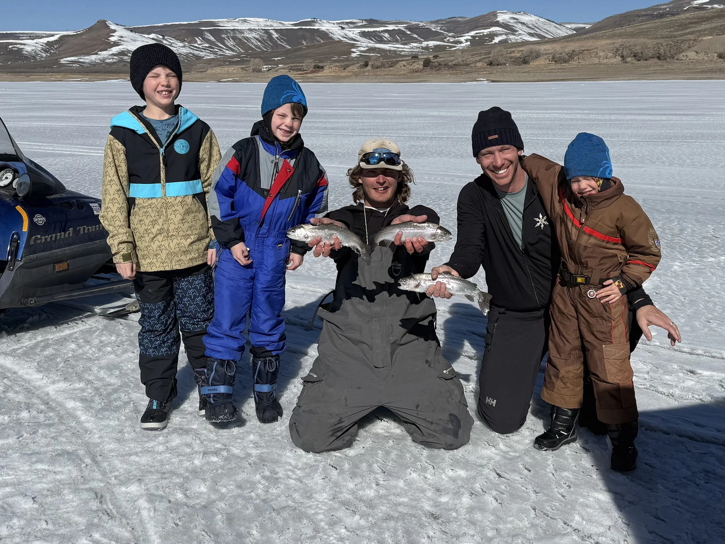 A family of six people, five children and one adult, standing on a snowy landscape holding fish. They are dressed in winter outdoor clothing, with a mountain and snow patches in the background. They are smiling and appear to have been fishing.