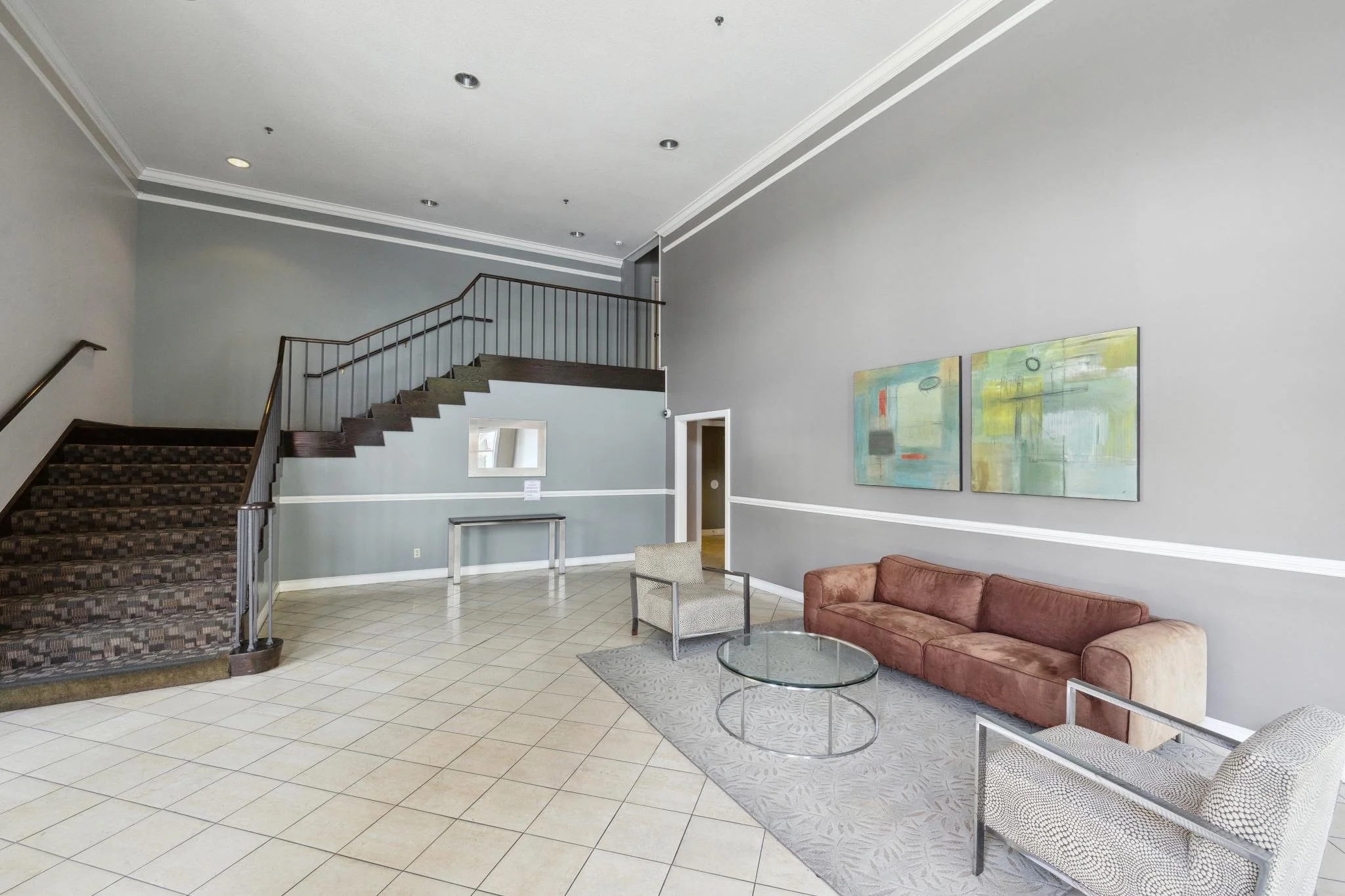 Interior of a modern lobby or waiting area with a staircase, light-colored tiled floor, gray walls with white trim, abstract paintings, a brown upholstered sofa, a round glass coffee table, and two chairs with metal frames and patterned cushions.