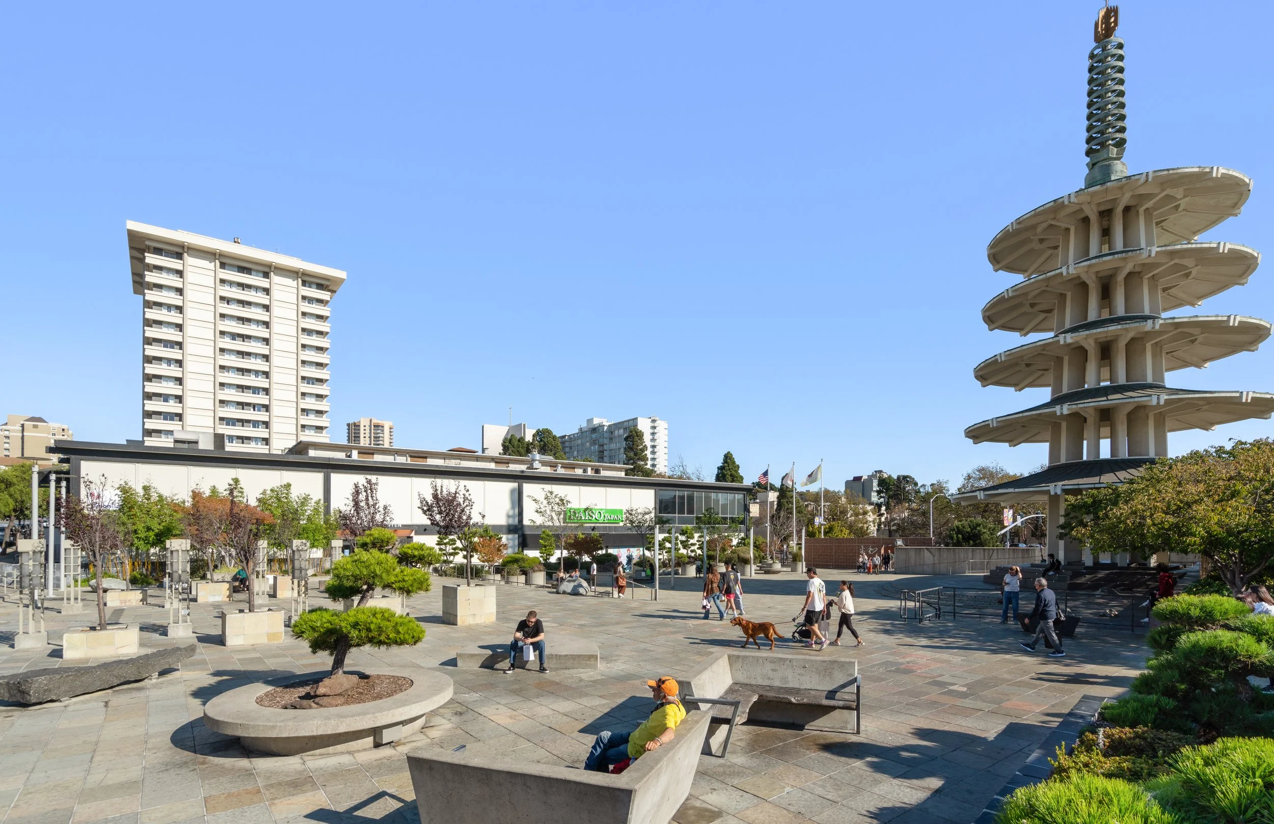 Urban park with people walking and sitting on benches, trees, and a tall pagoda structure under a clear blue sky with high-rise buildings in the background.