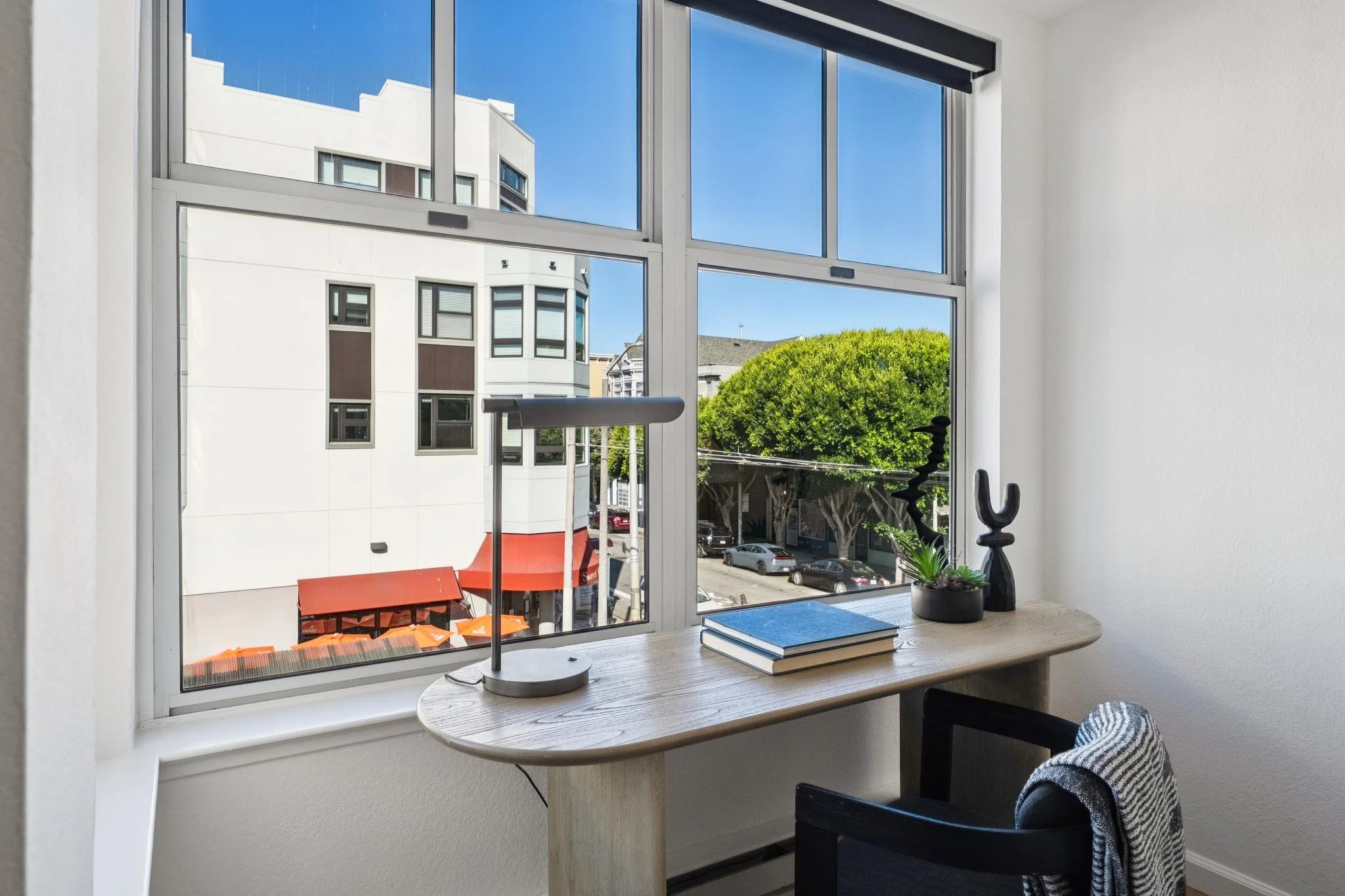 A view through a large window showing a city street with parked cars, trees, and modern buildings. Inside, a wooden desk with books, a black sculpture, a small plant, and a desk lamp are visible, with part of a chair and a striped chair cushion.